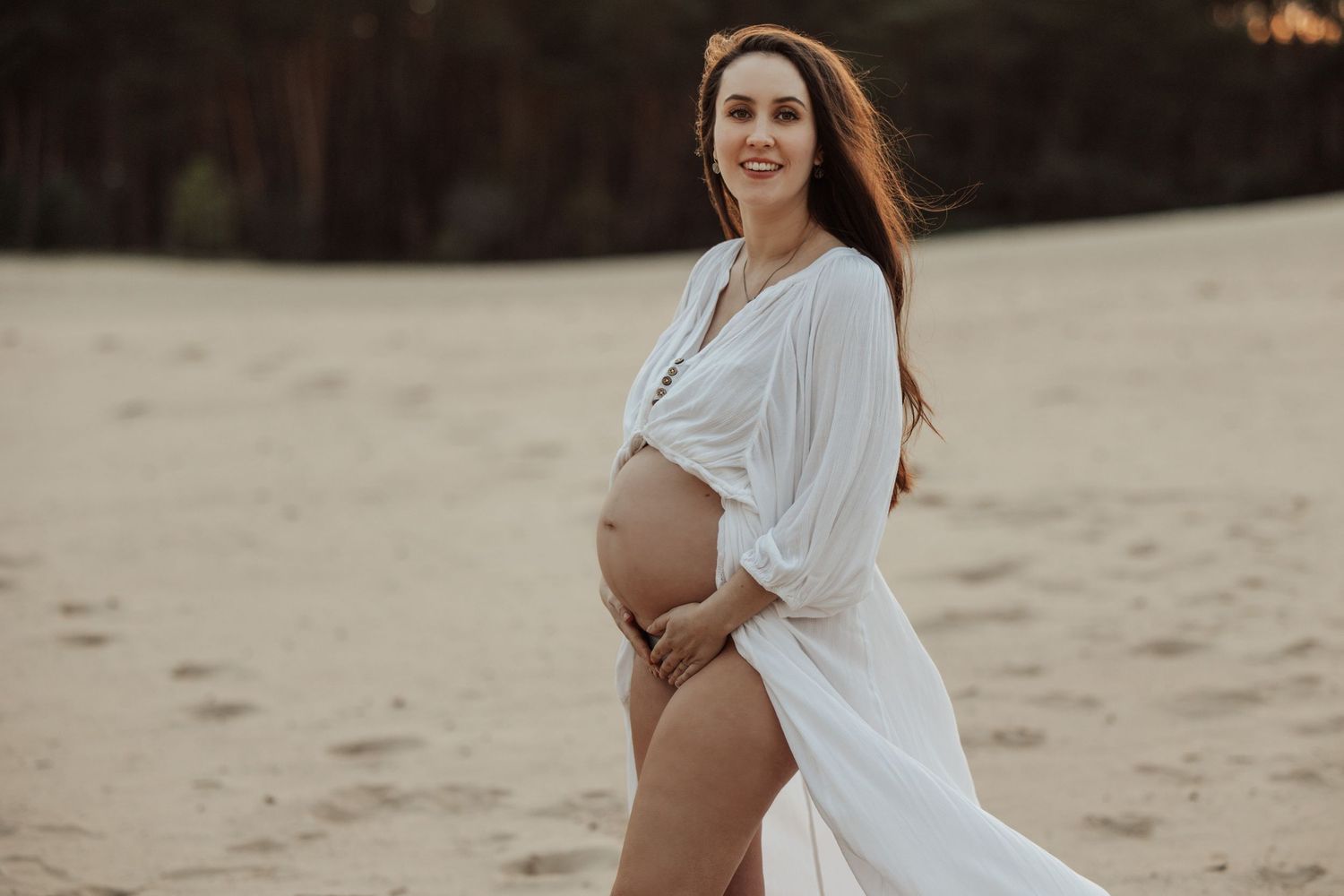 Ethereal maternity portraits in flowing white dress during sunset beach session.