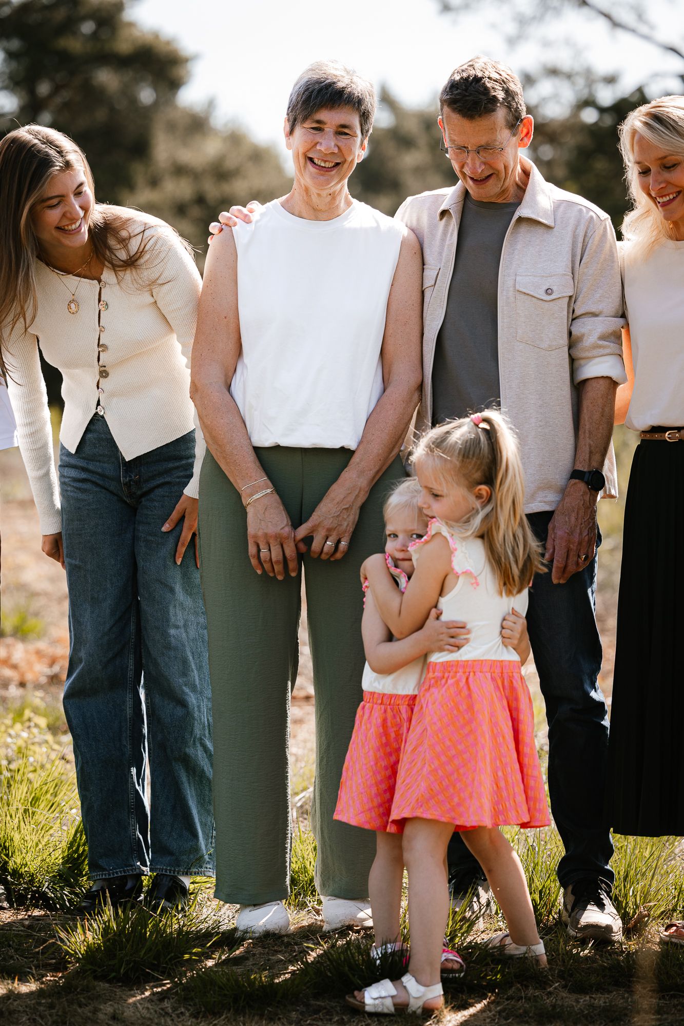 Multi-generational family smiling outdoors, with two young girls hugging in front of grandparents and adult women.