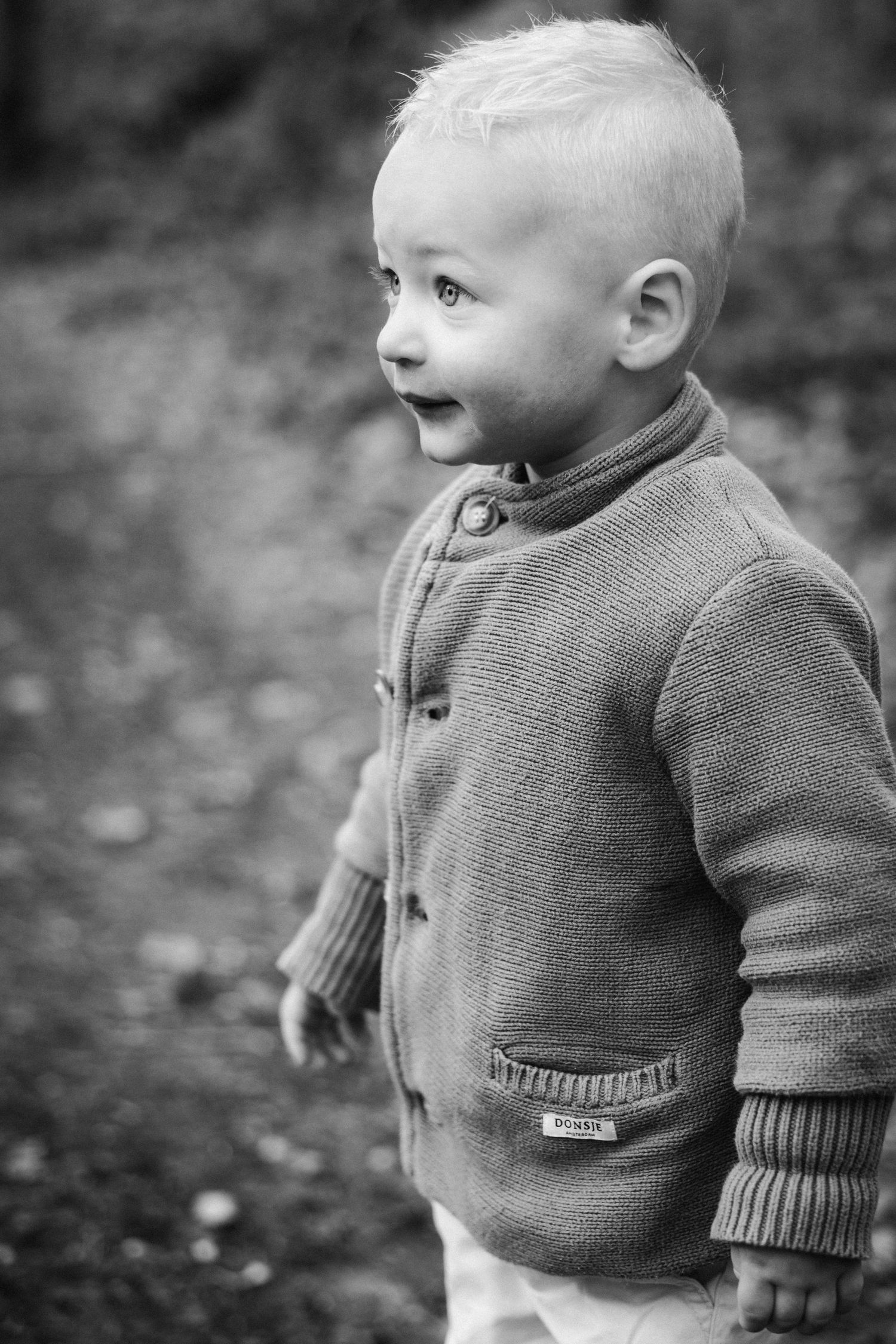 Black and white portrait of a curious toddler boy in a knit cardigan sweater standing outdoors.