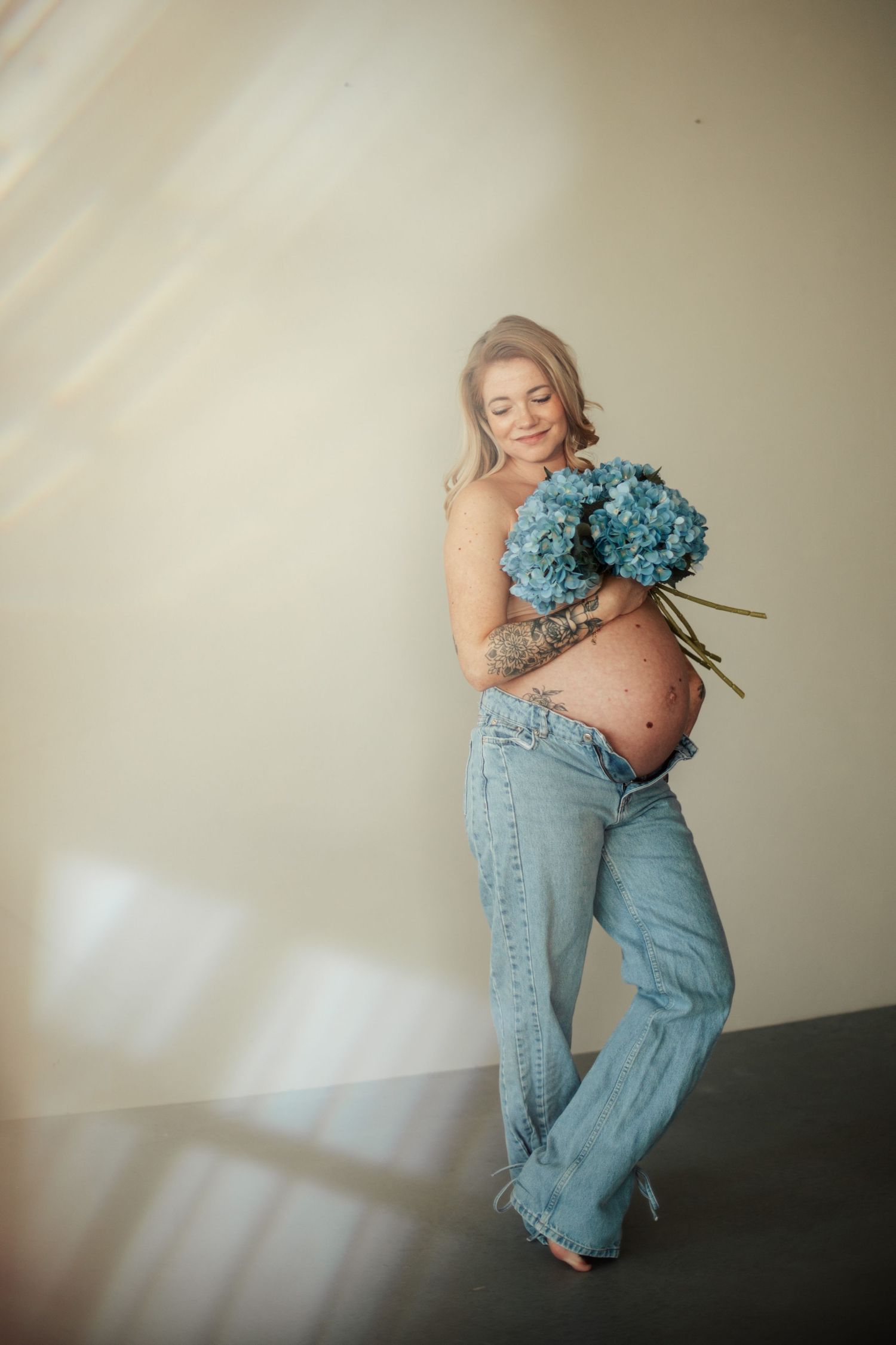 Maternity photo with woman in jeans holding blue hydrangeas against soft beige background.