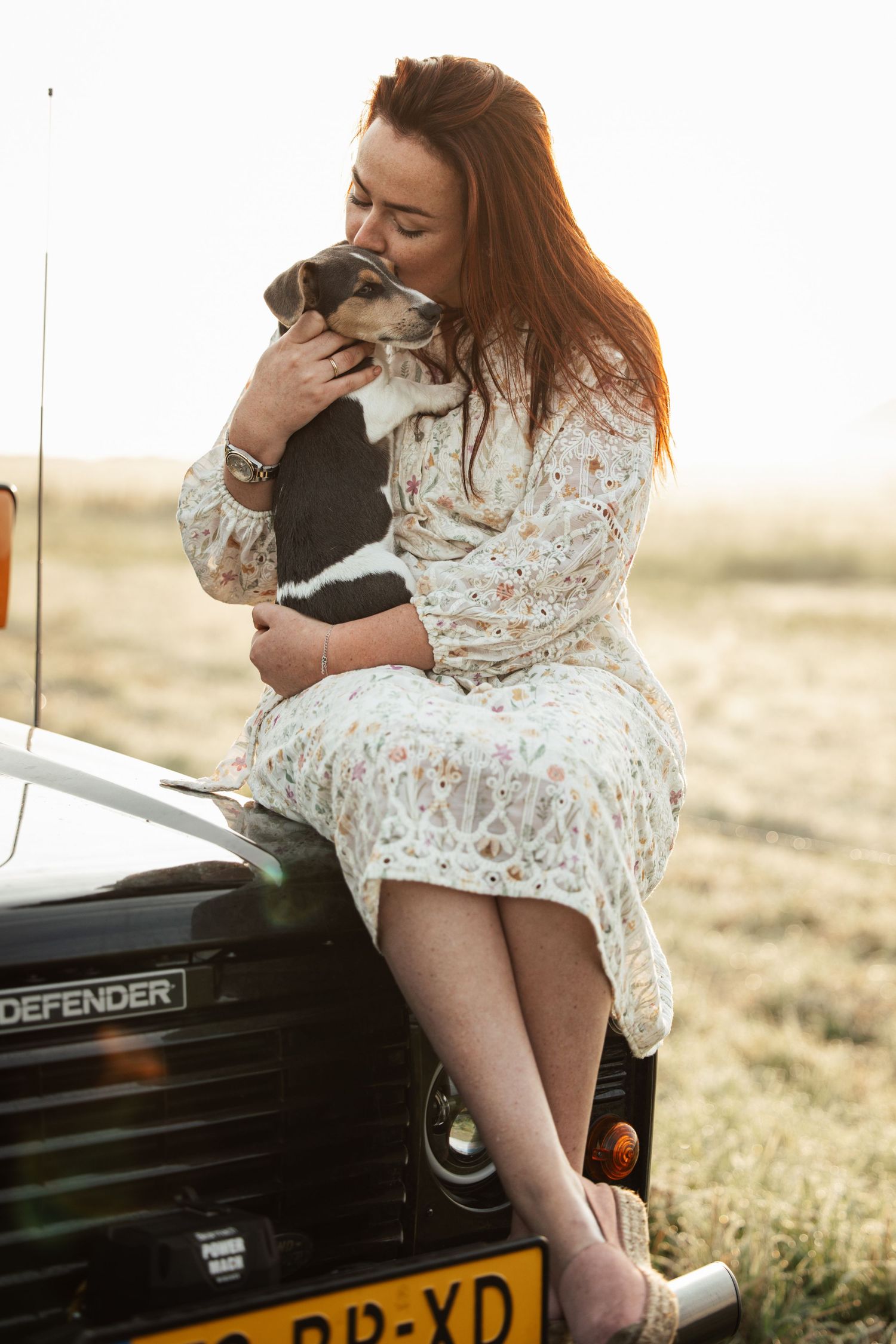 Person in white lace dress sitting on black Land Defender vehicle holding small dog in countryside setting.