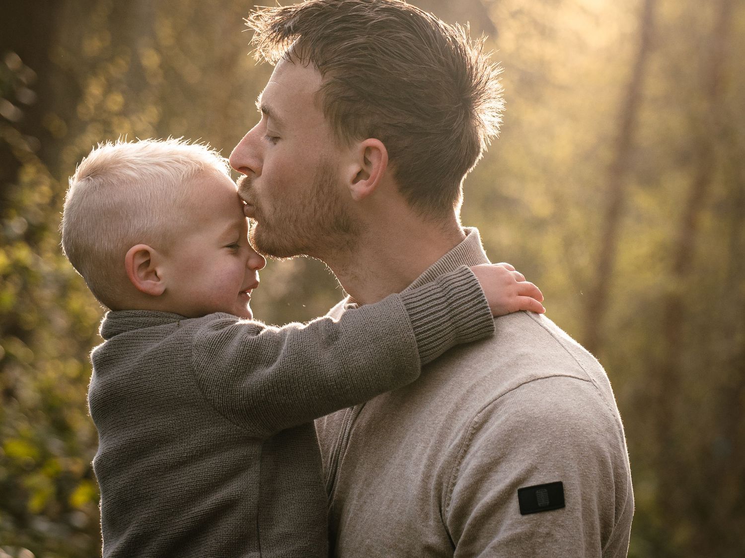 A father tenderly kisses his young toddler son outdoors in warm golden sunlight among green trees.