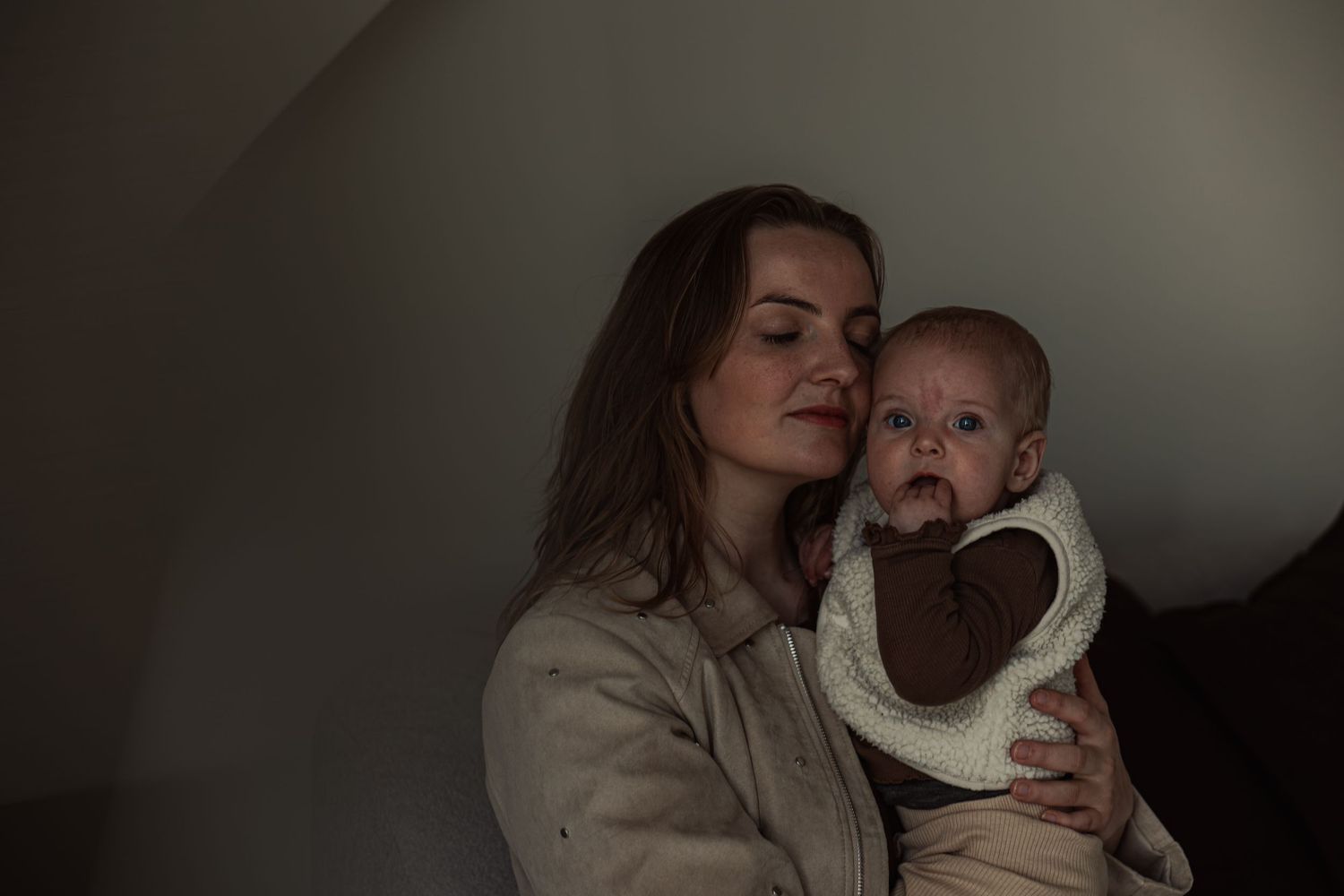 A young mother tenderly holds her alert baby, both dressed in neutral tones against a softly lit background.
