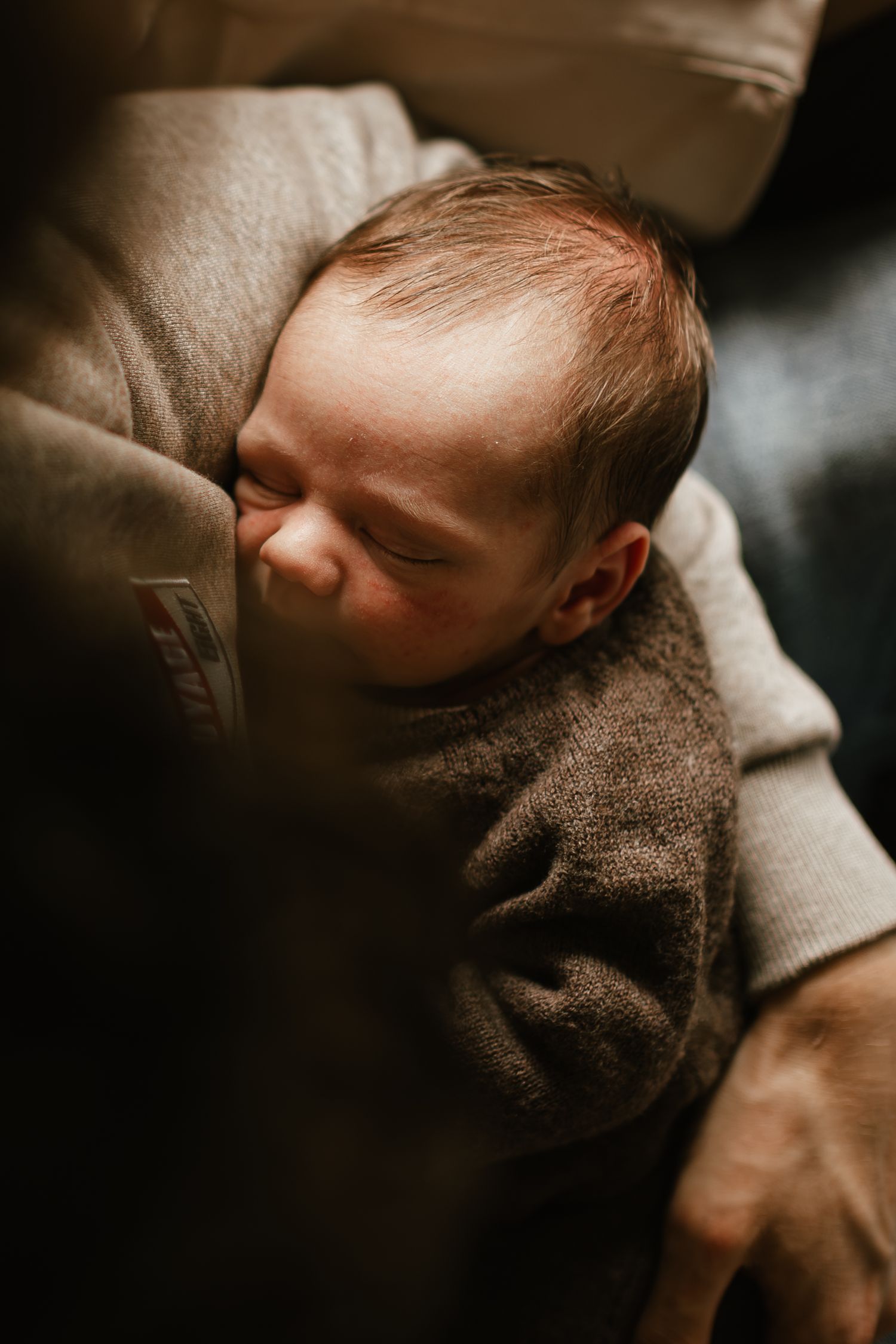 Newborn baby sleeping peacefully in a parent's arms, wrapped in a soft brown knit sweater.