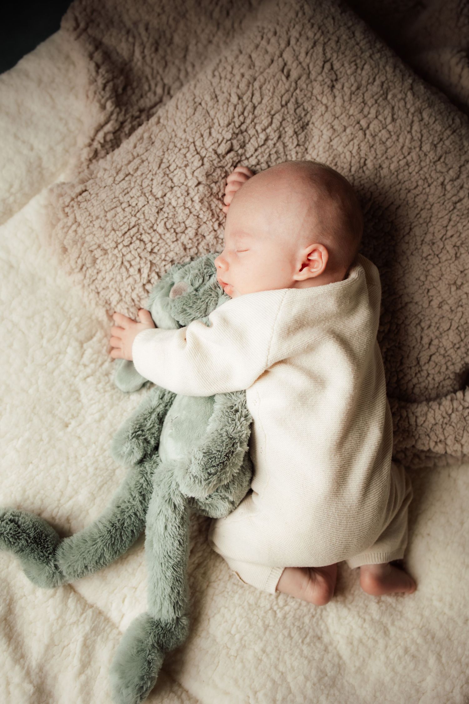 Baby in white sweater lying on carpet with soft green plush toy.