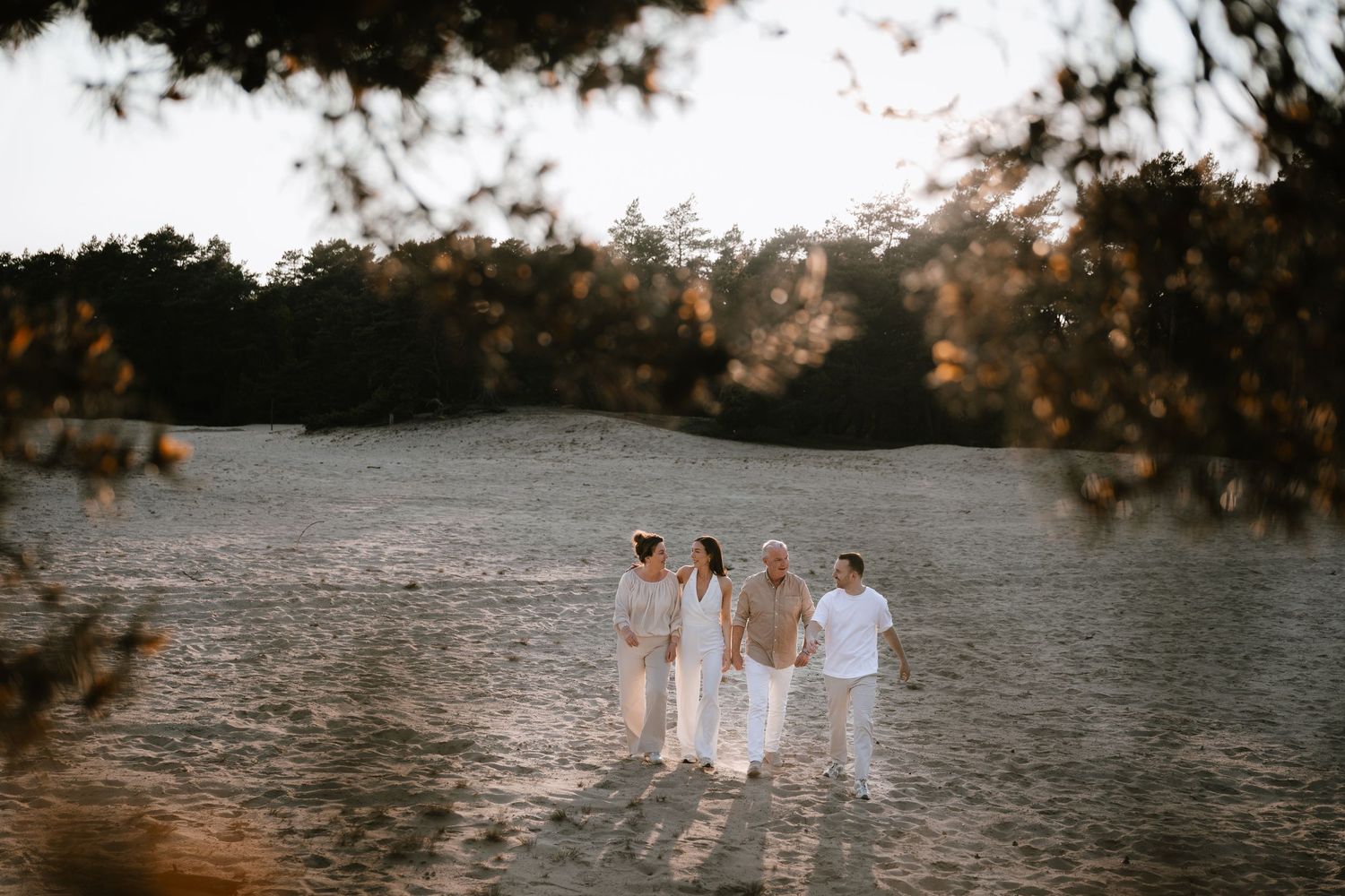 Four family members walking together on golden sandy dunes surrounded by trees at sunset.