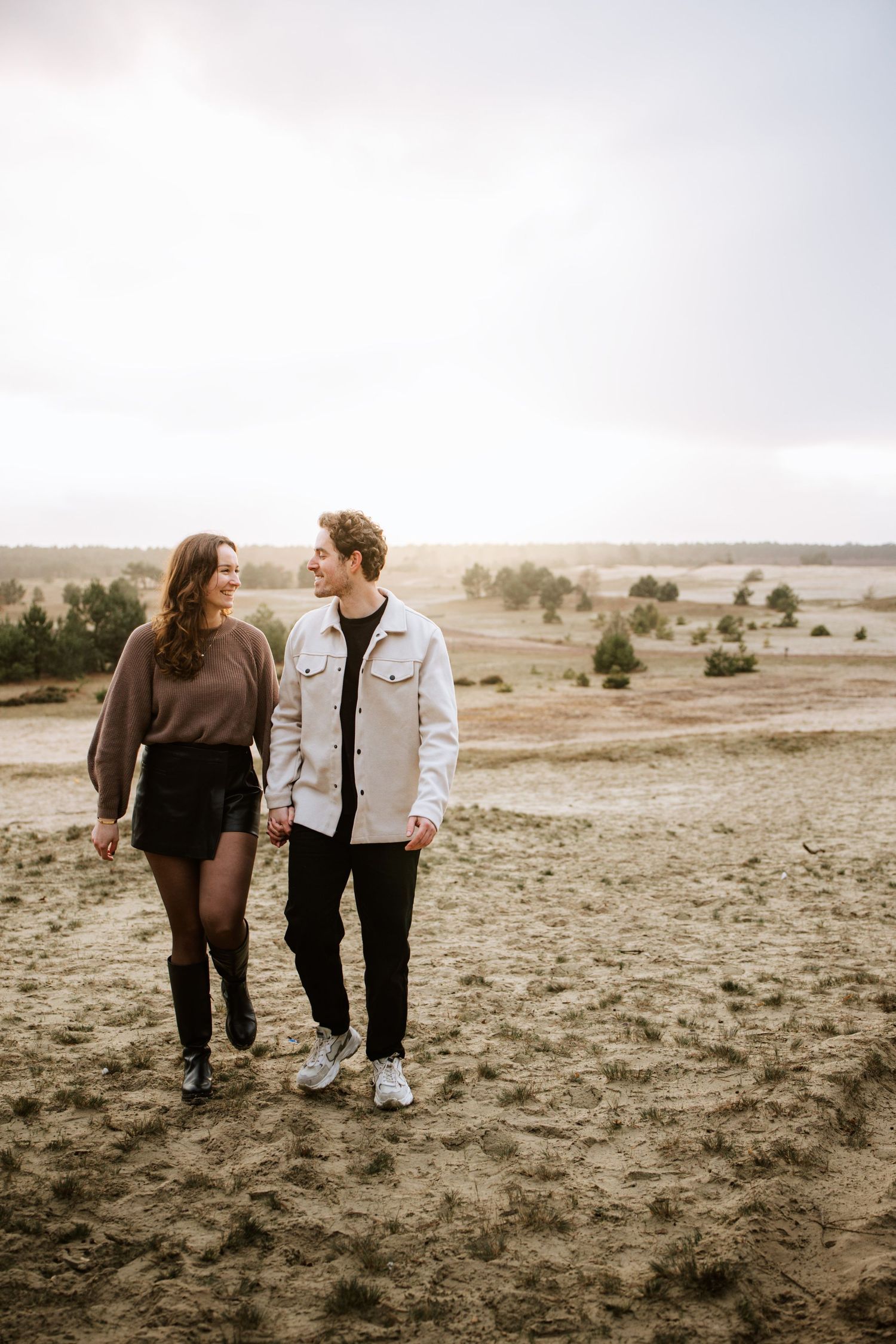 A couple walks hand in hand across a barren landscape at sunset.