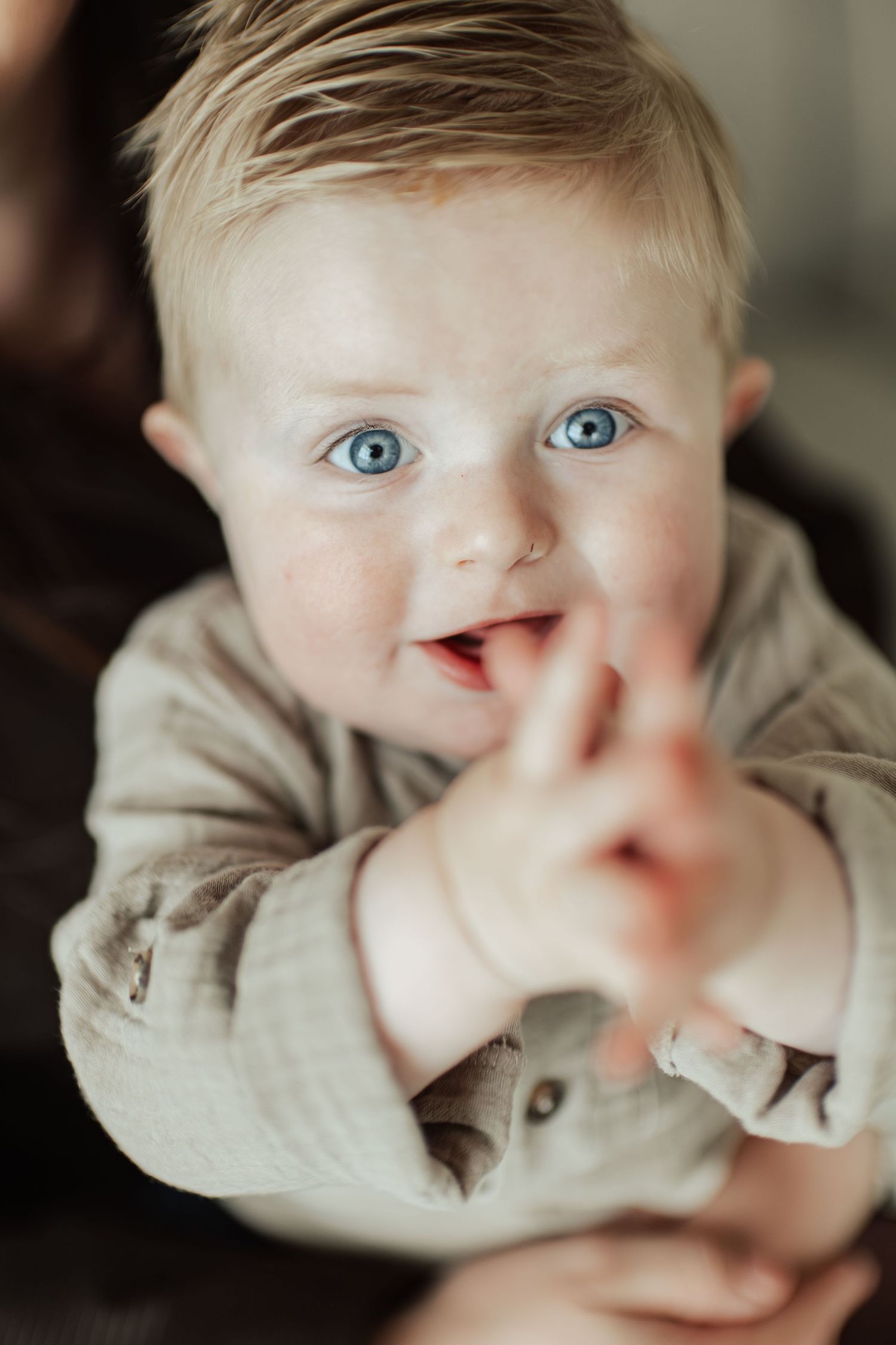 Adorable blue-eyed baby with blonde hair smiling and reaching toward camera in a cozy brown outfit.