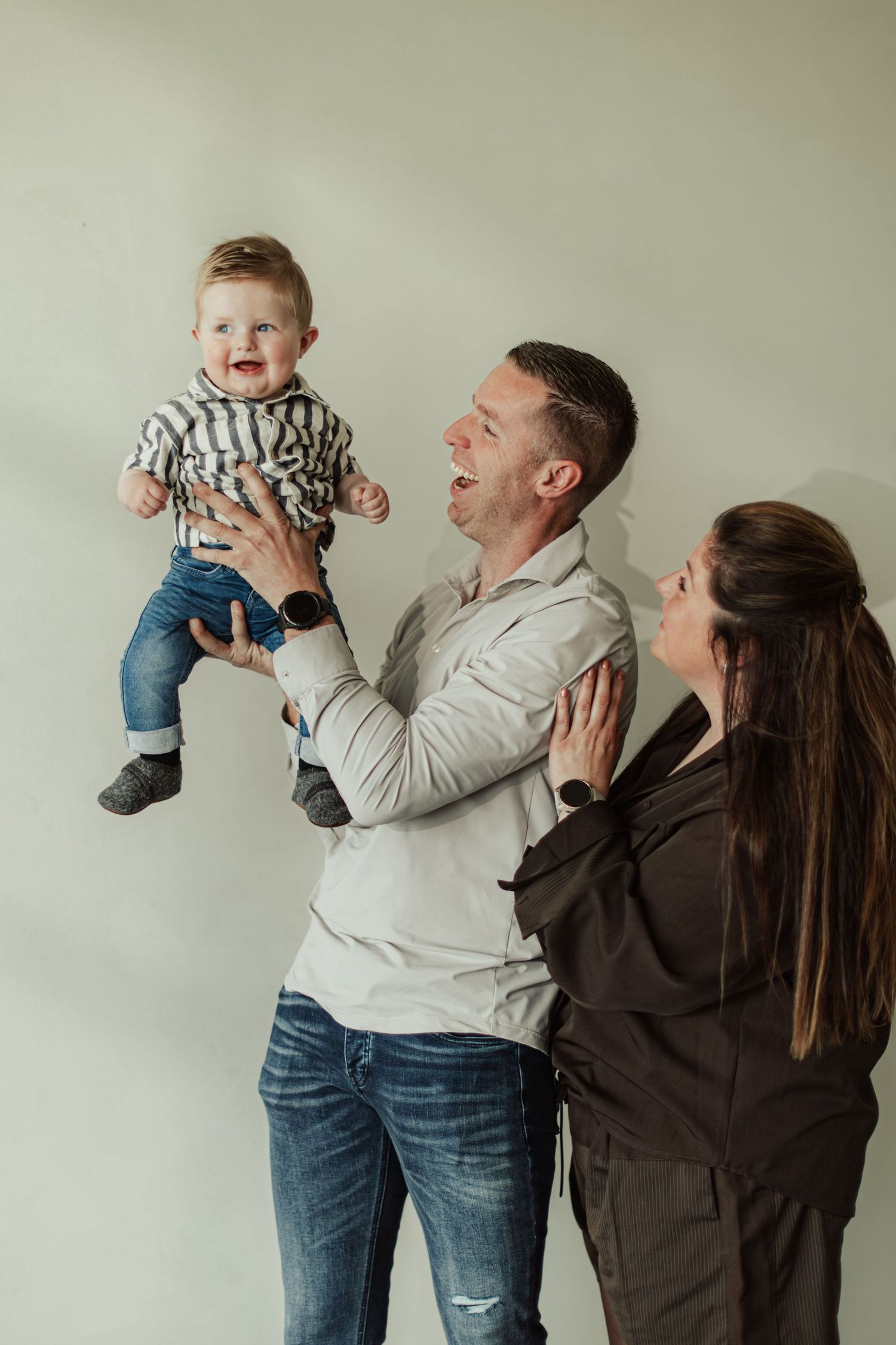 Happy family with father lifting smiling baby boy while mother watches joyfully against light background.