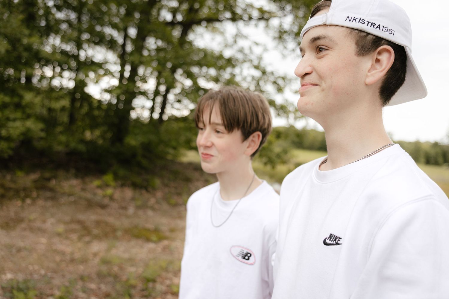 Two young people in white Nike and New Balance shirts standing outdoors near trees on a sunny day.