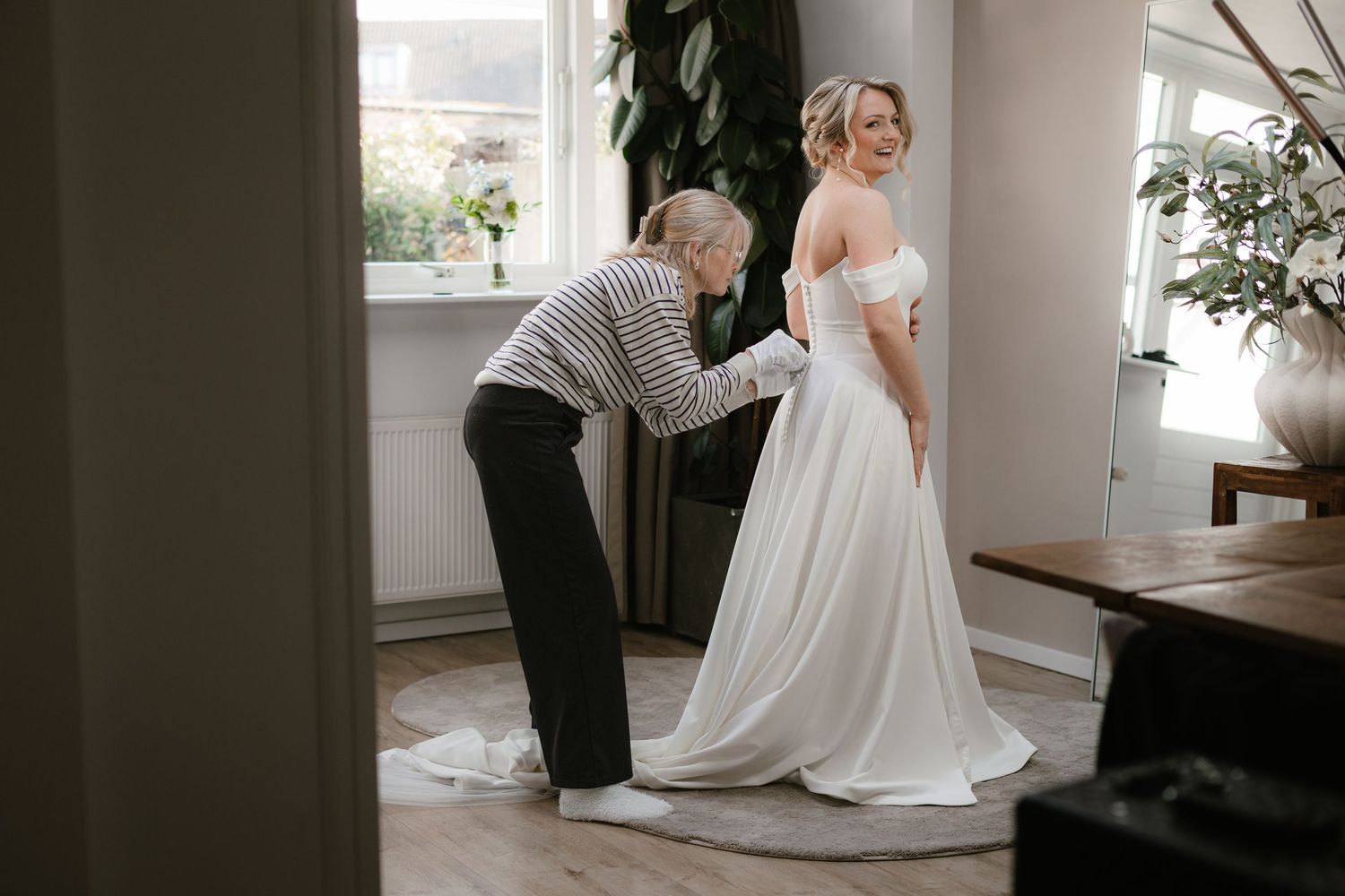 A seamstress adjusts the back of a brides elegant white off-shoulder wedding gown in a bright, stylish room.
