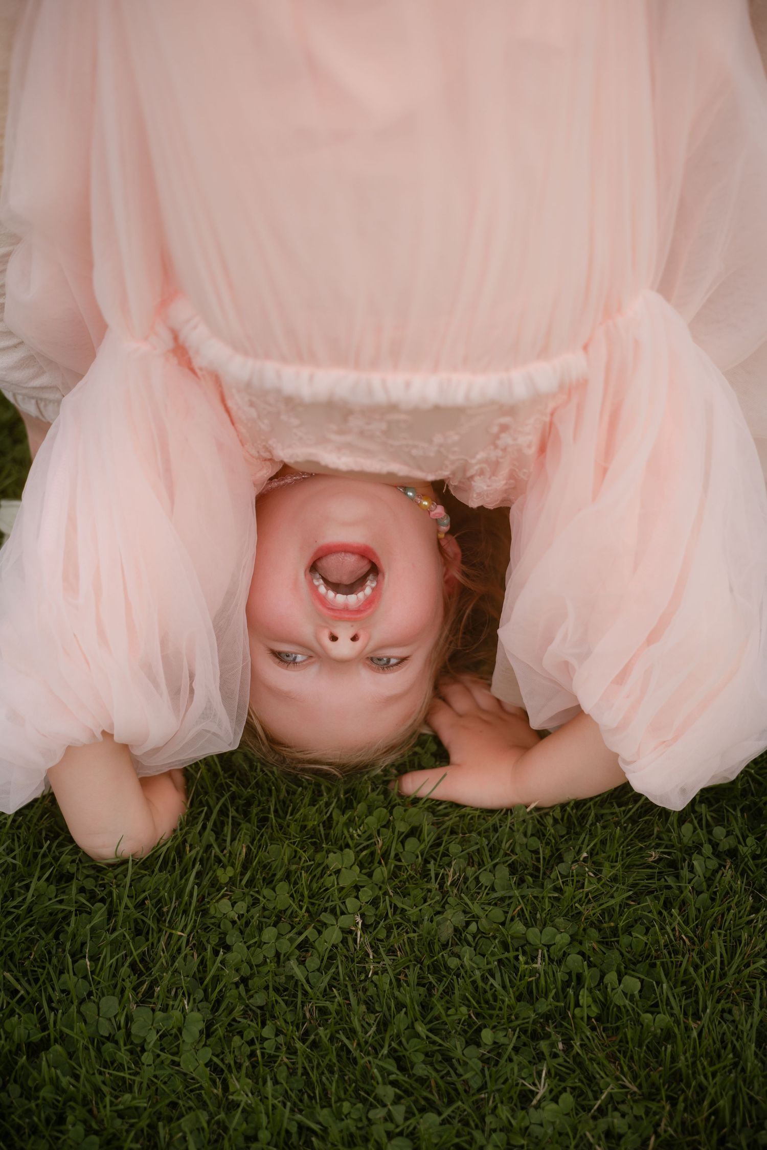 Toddler girl in pink dress bending over upside down on green grass with a pacifier in her mouth.