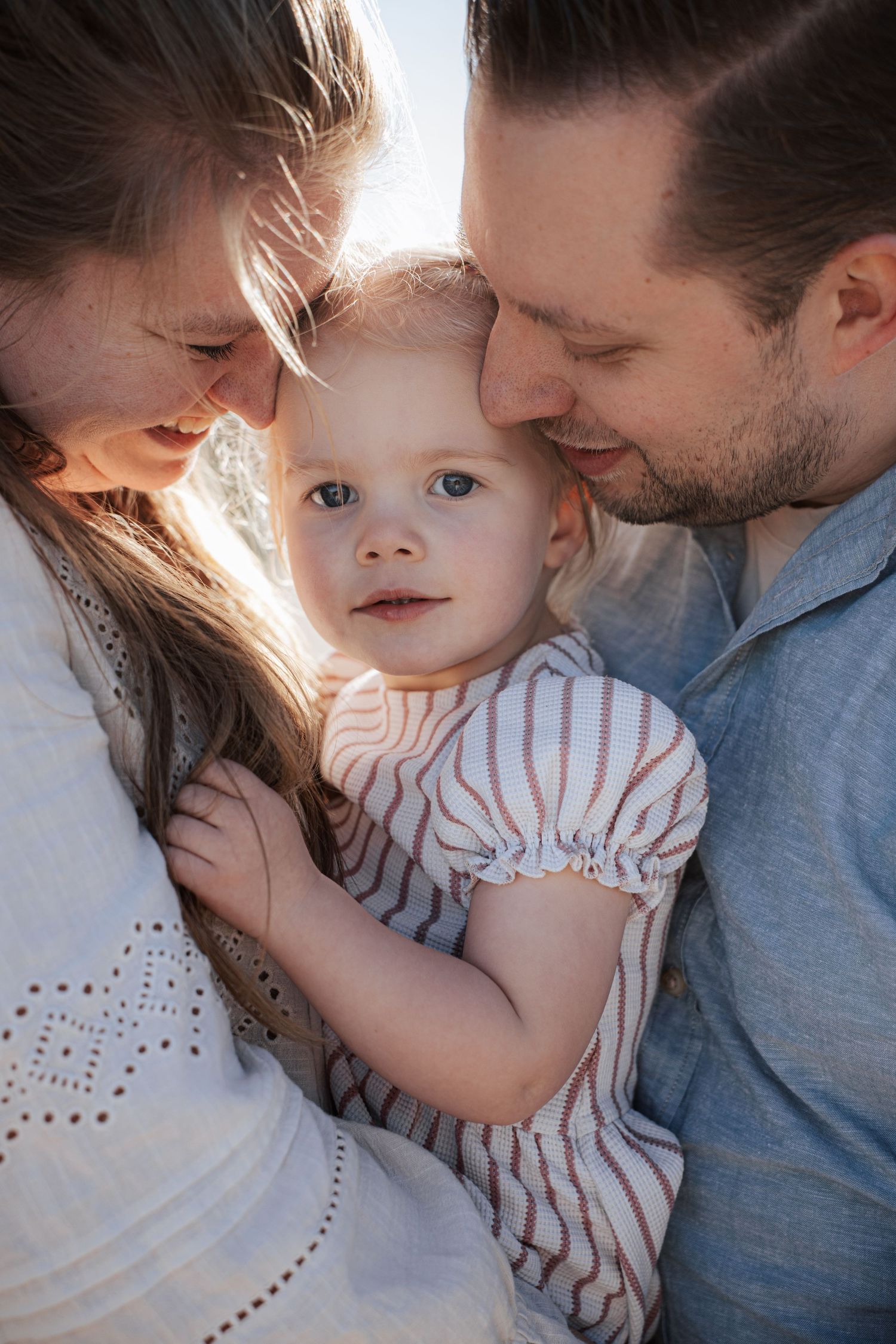 A young family with parents lovingly nuzzling their smiling baby girl in warm golden sunlight outdoors.