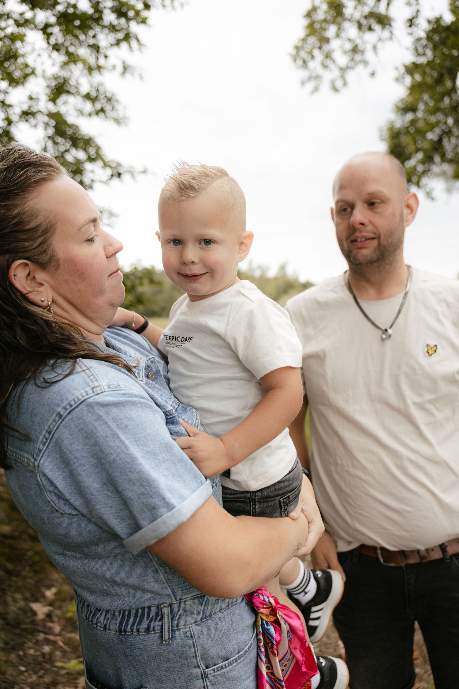 A woman holds a young blonde toddler boy while a bald man stands beside them outdoors near trees.