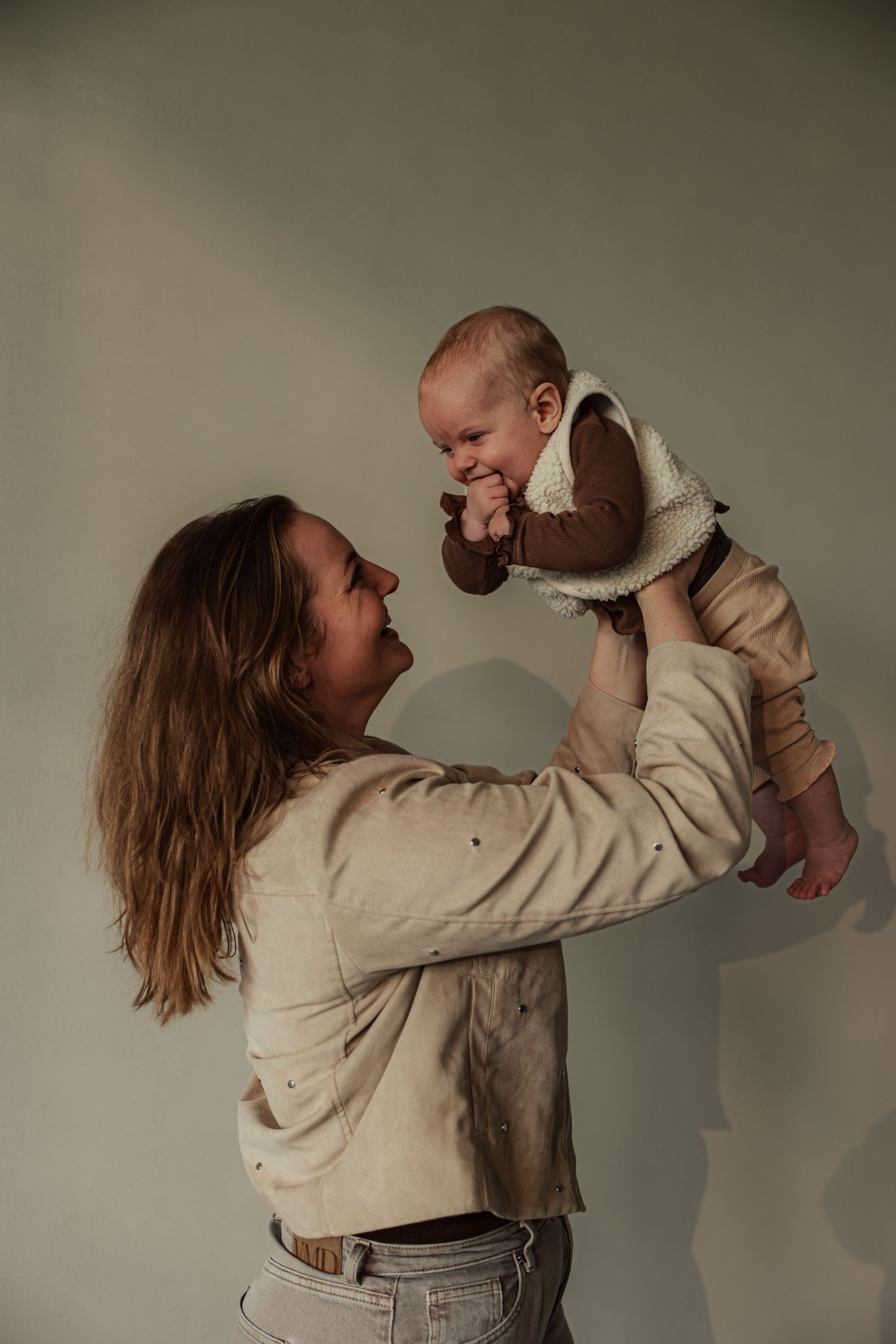 A joyful mother lifts her smiling baby in the air against a soft green background, both wearing neutral tones.