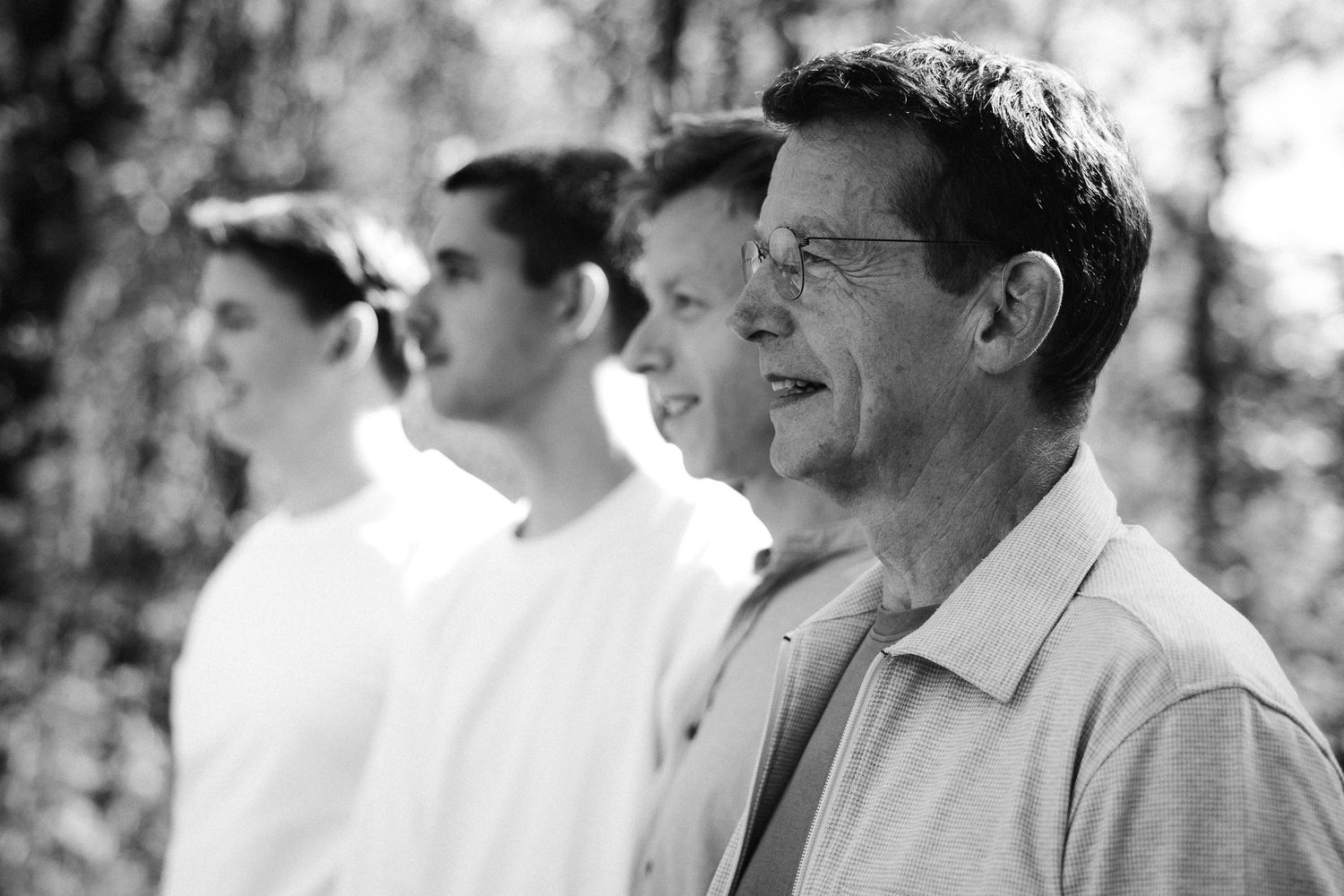 Black and white photo of four men standing in a row outdoors among trees, viewed from the side.