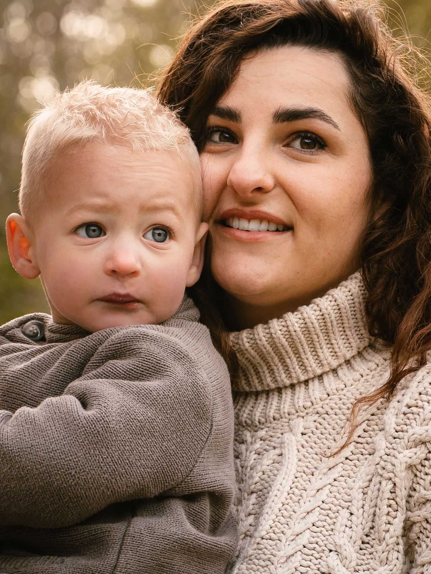 A smiling brunette woman in a cream cable-knit sweater holds a blue-eyed toddler boy outdoors in soft natural light.