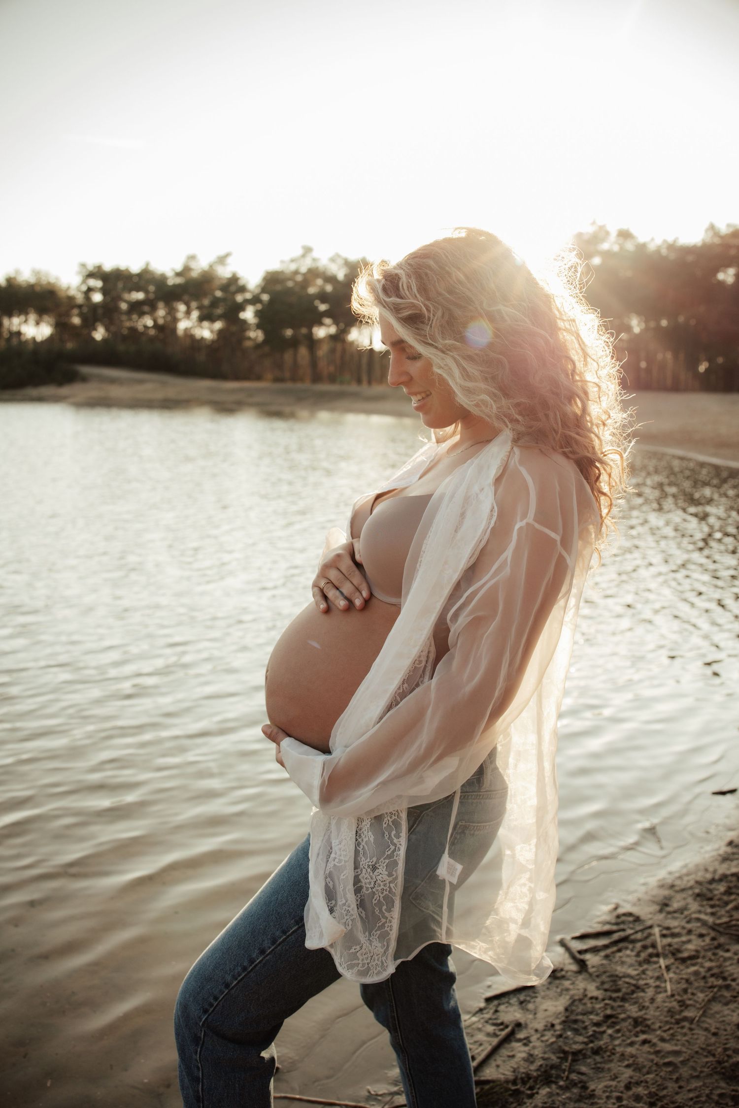 Maternity photo session at sunset by a lake with flowing white dress and natural lighting.