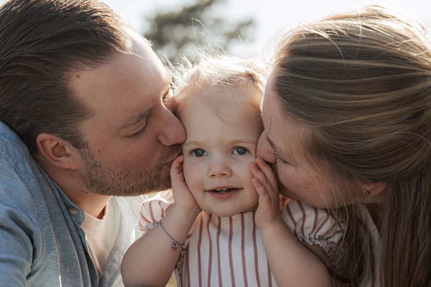 Two parents kissing their smiling baby on the cheeks in a warm, sunny outdoor family portrait.