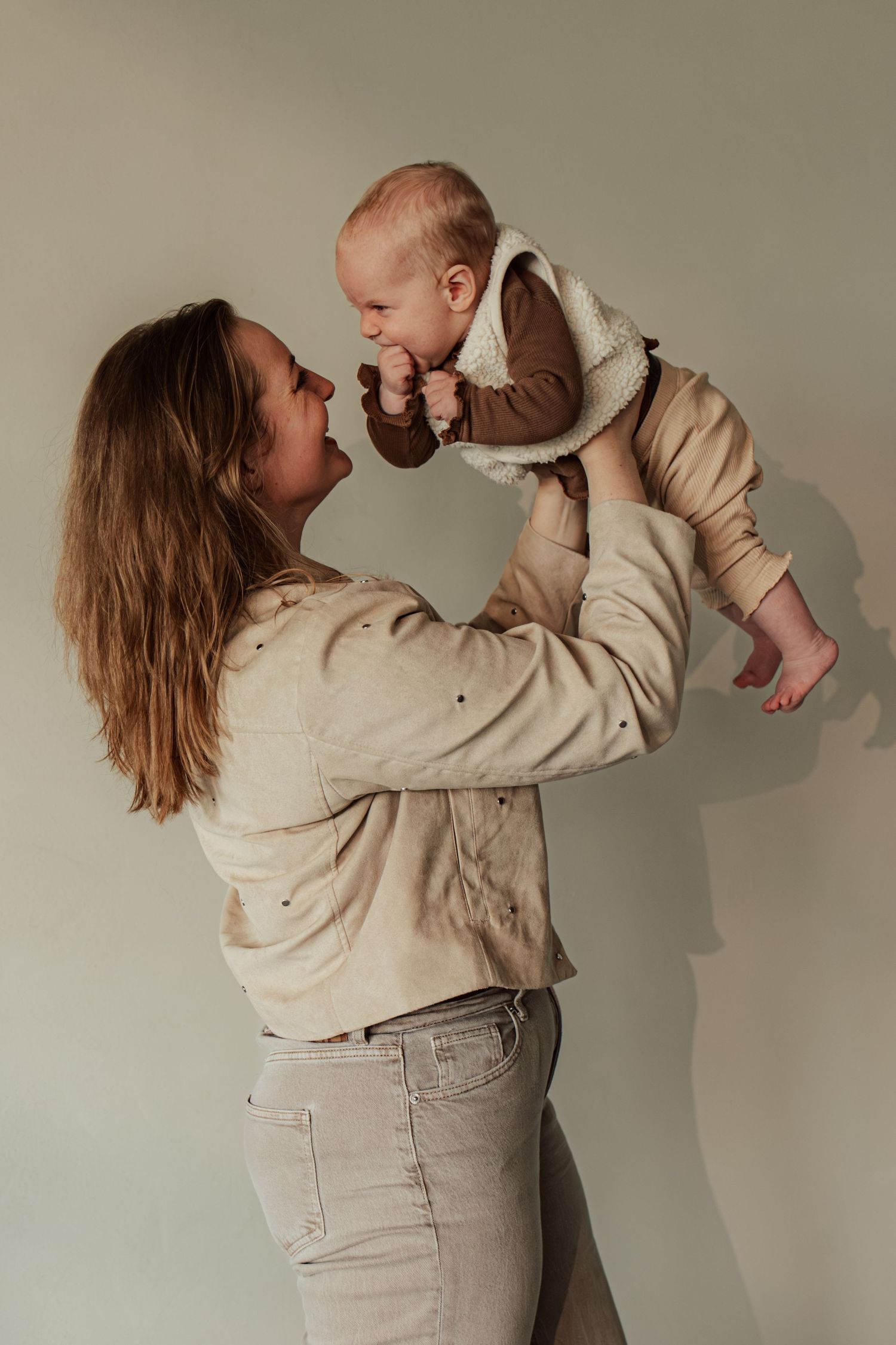 A mother in beige jacket lifts her smiling baby wearing a brown vest, sharing a joyful moment against a light background.