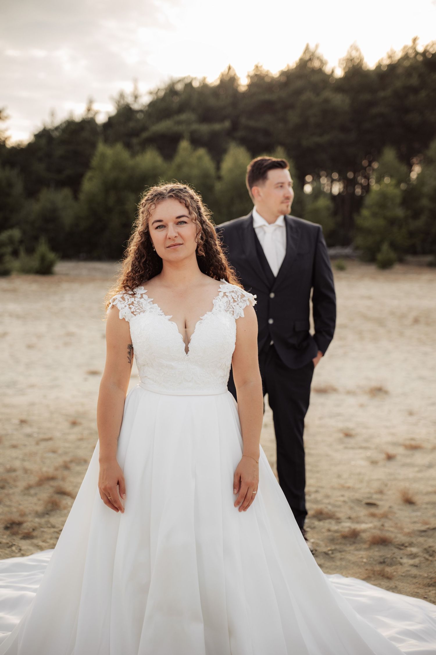 Bride wearing lace wedding gown poses in natural outdoor setting.