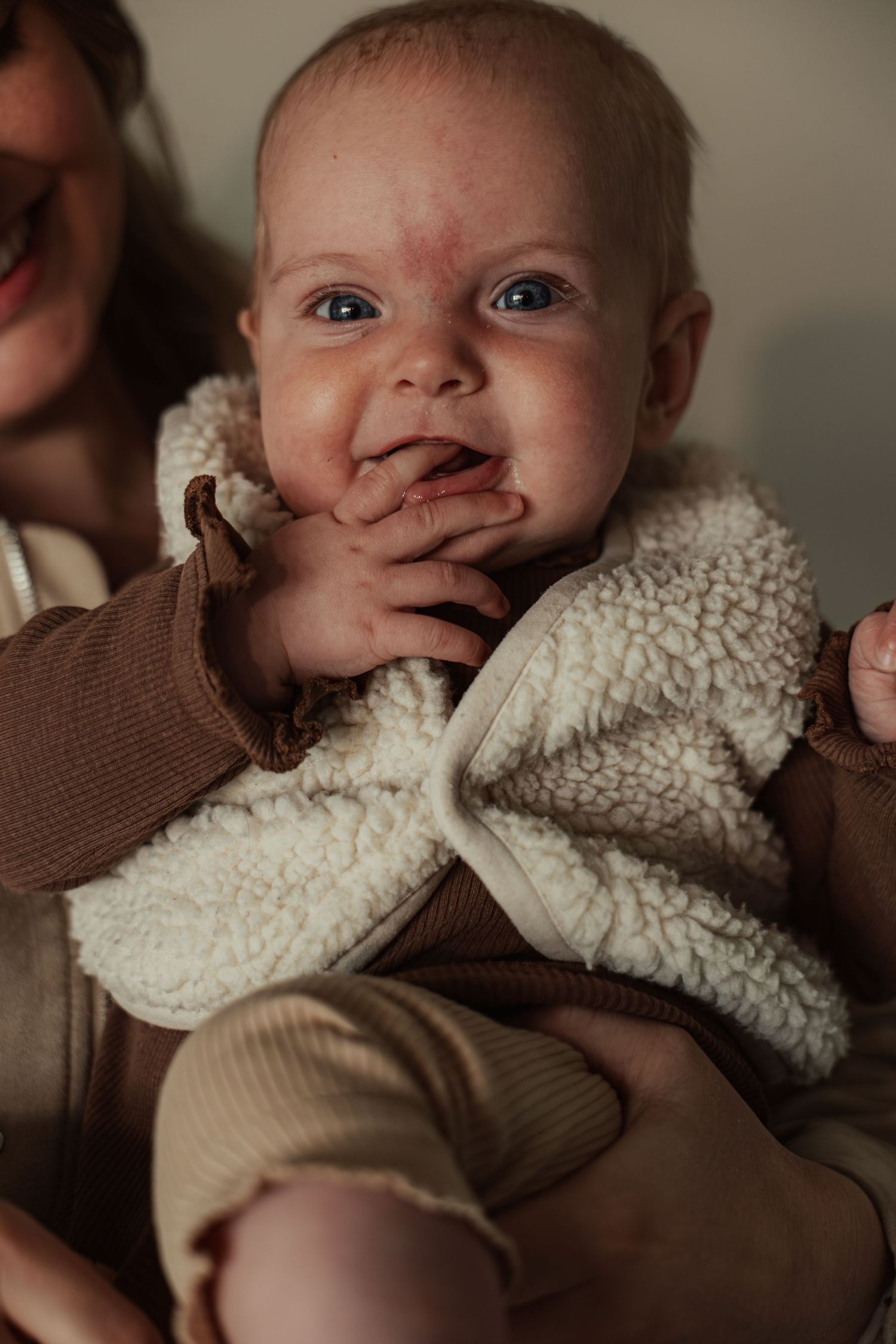 Adorable baby in white sherpa vest smiling with hand near mouth, held by mother in cozy indoor setting.