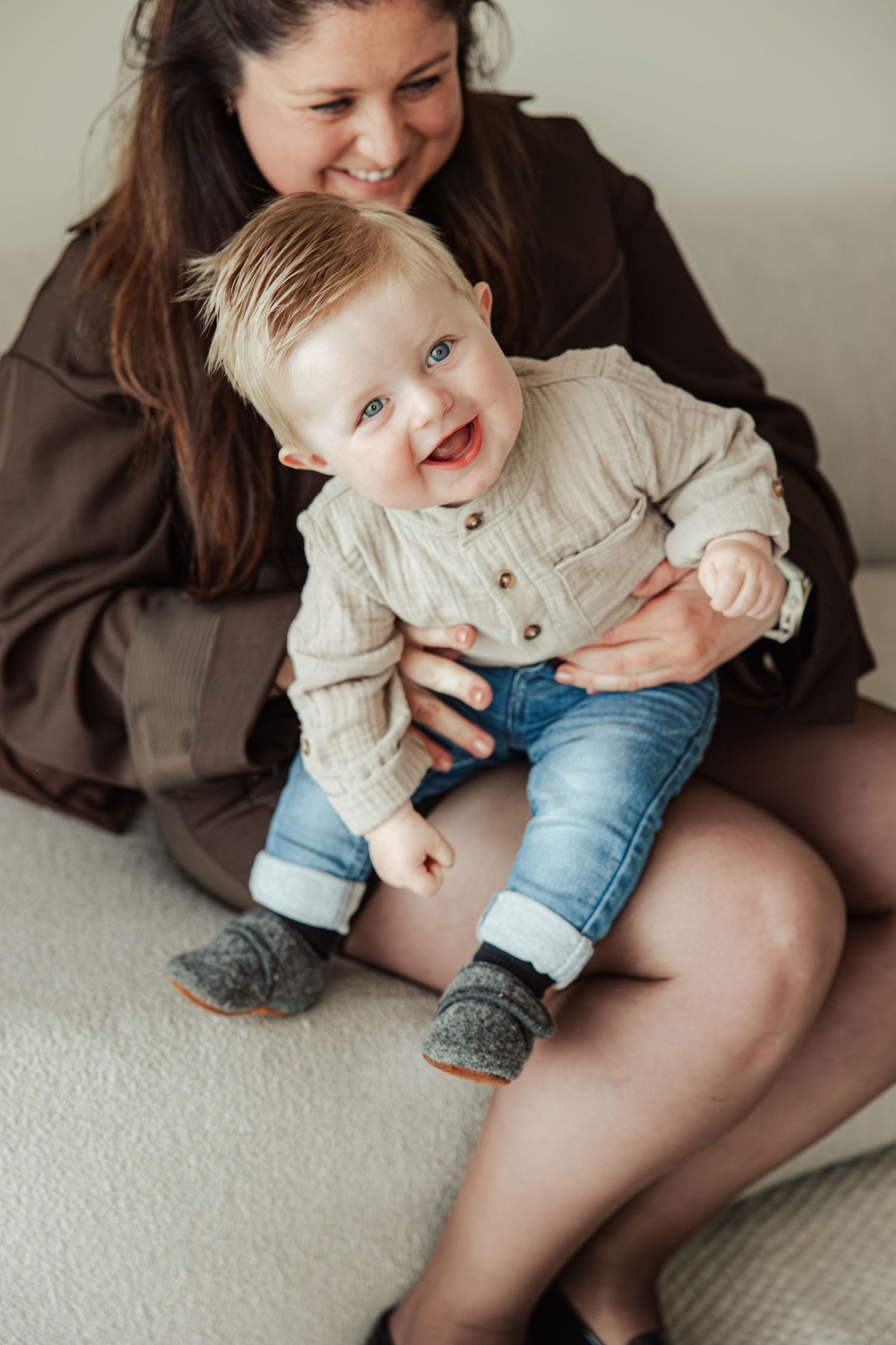 A joyful baby boy in a beige sweater and jeans laughs while sitting on his smiling mother's lap indoors.