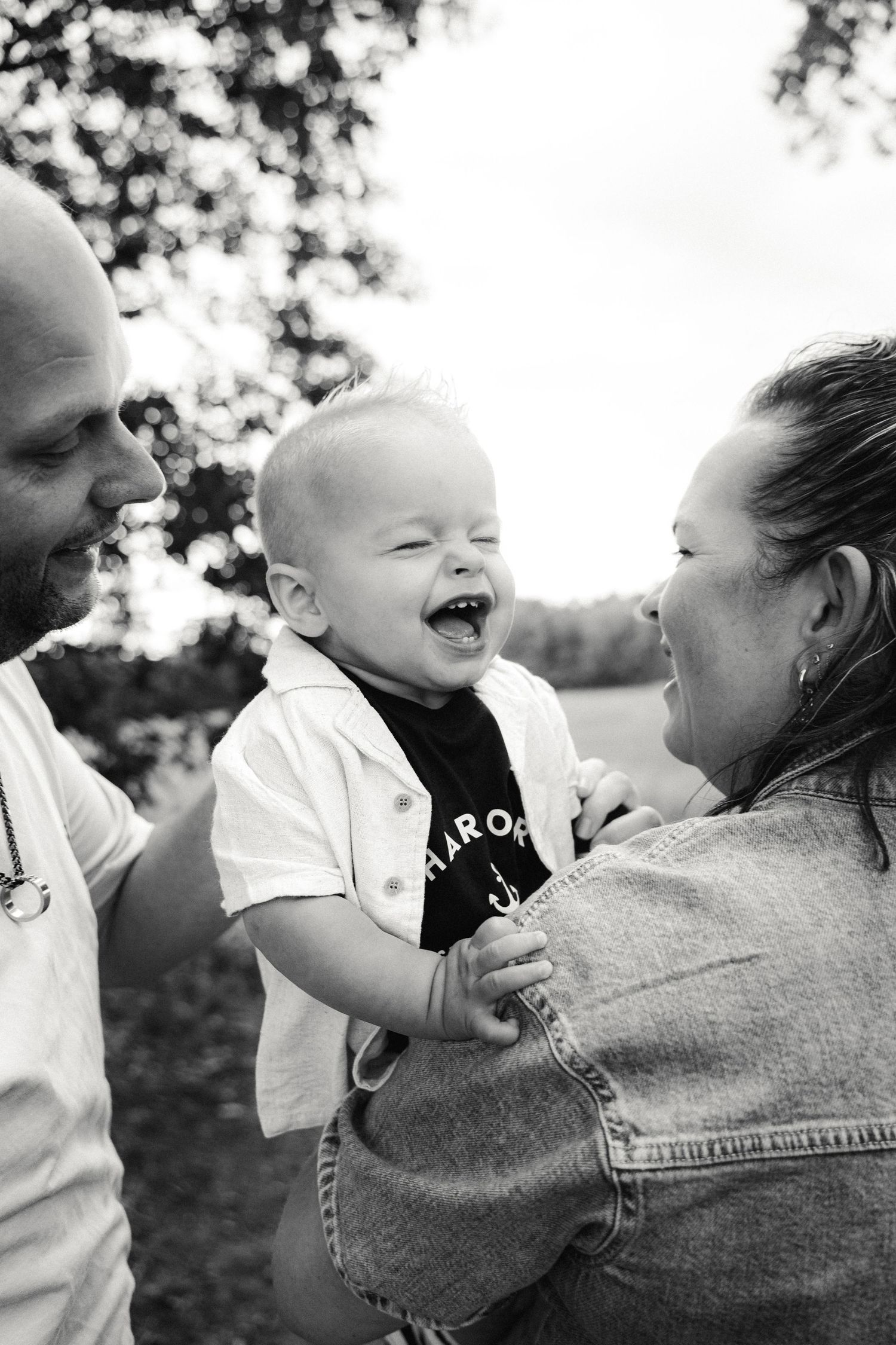 Black and white photo of laughing baby held by smiling parents outdoors near trees.