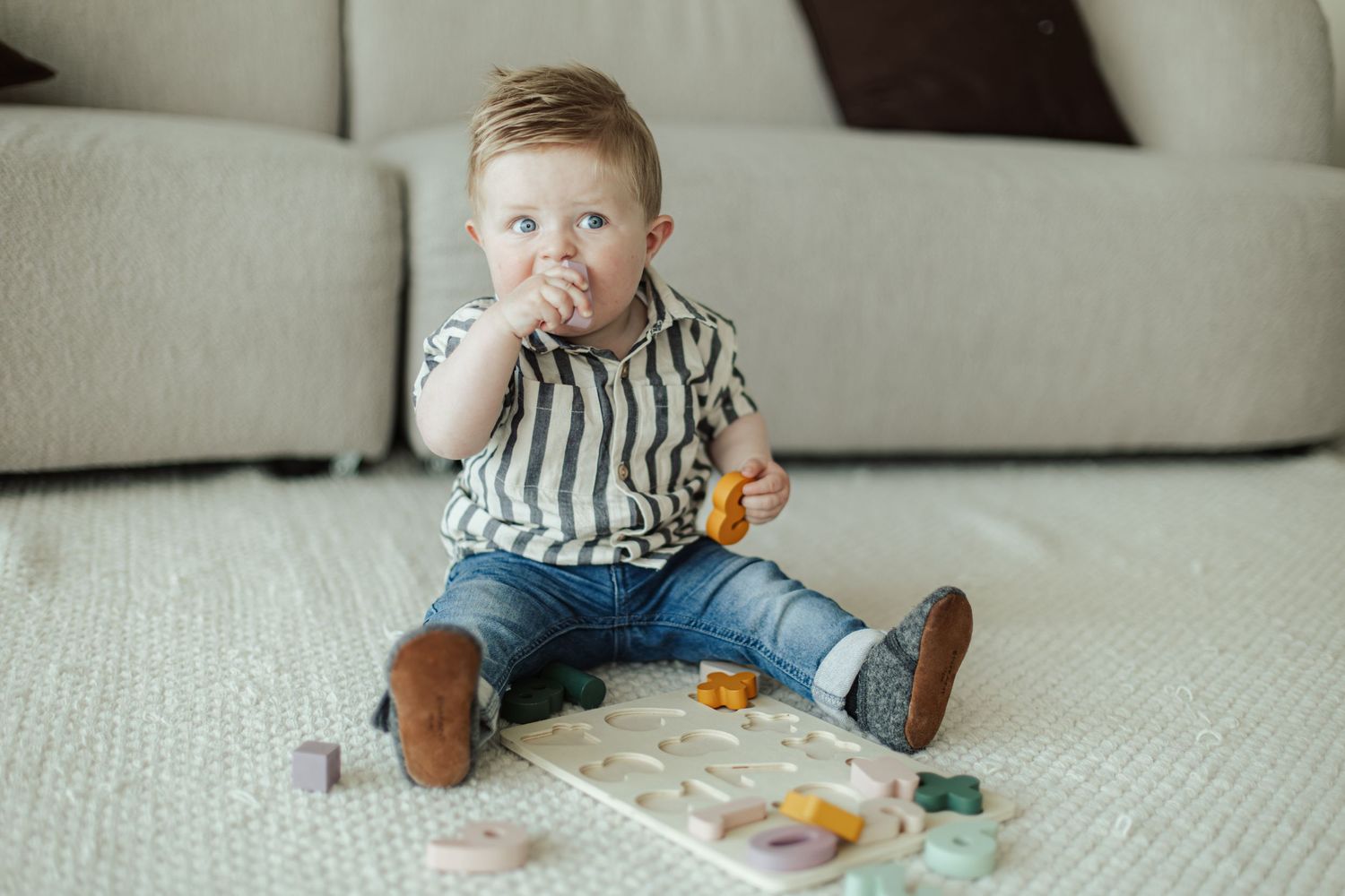 Cute toddler boy in striped shirt and jeans sitting on carpet floor playing with colorful wooden toys.