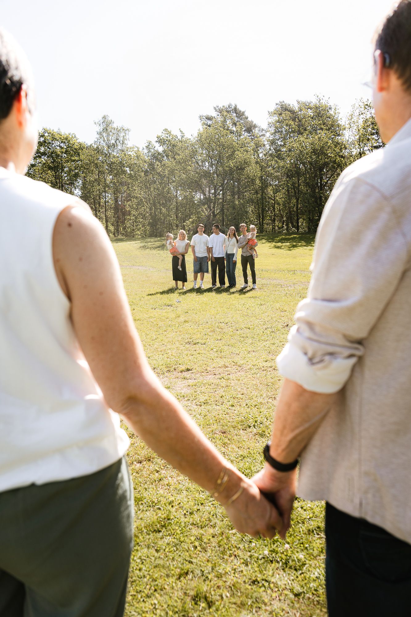 Two people holding hands walk toward a group of friends gathered on a sunny green lawn surrounded by trees.