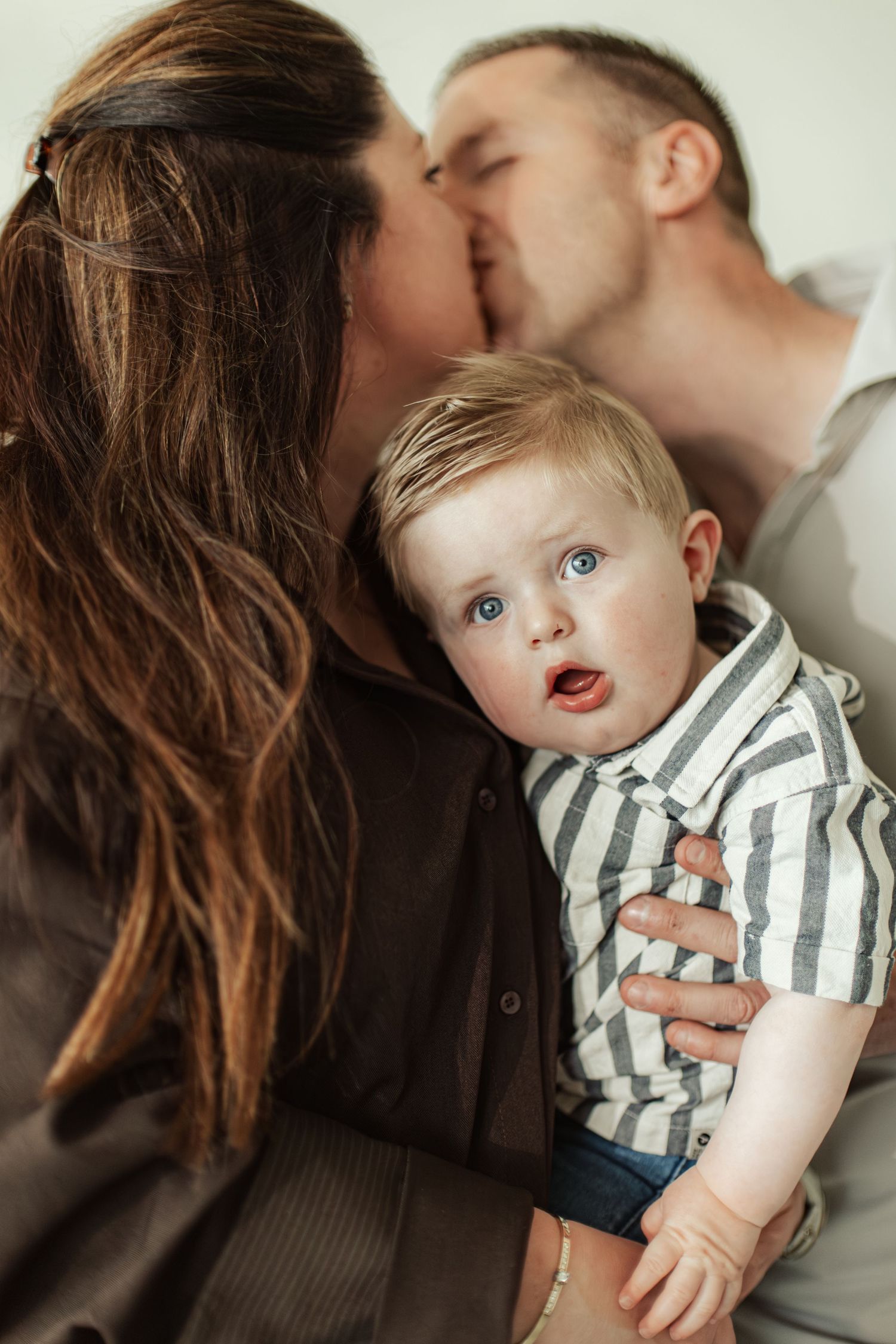 A couple kissing behind their wide-eyed baby boy in a striped shirt during a family portrait session.