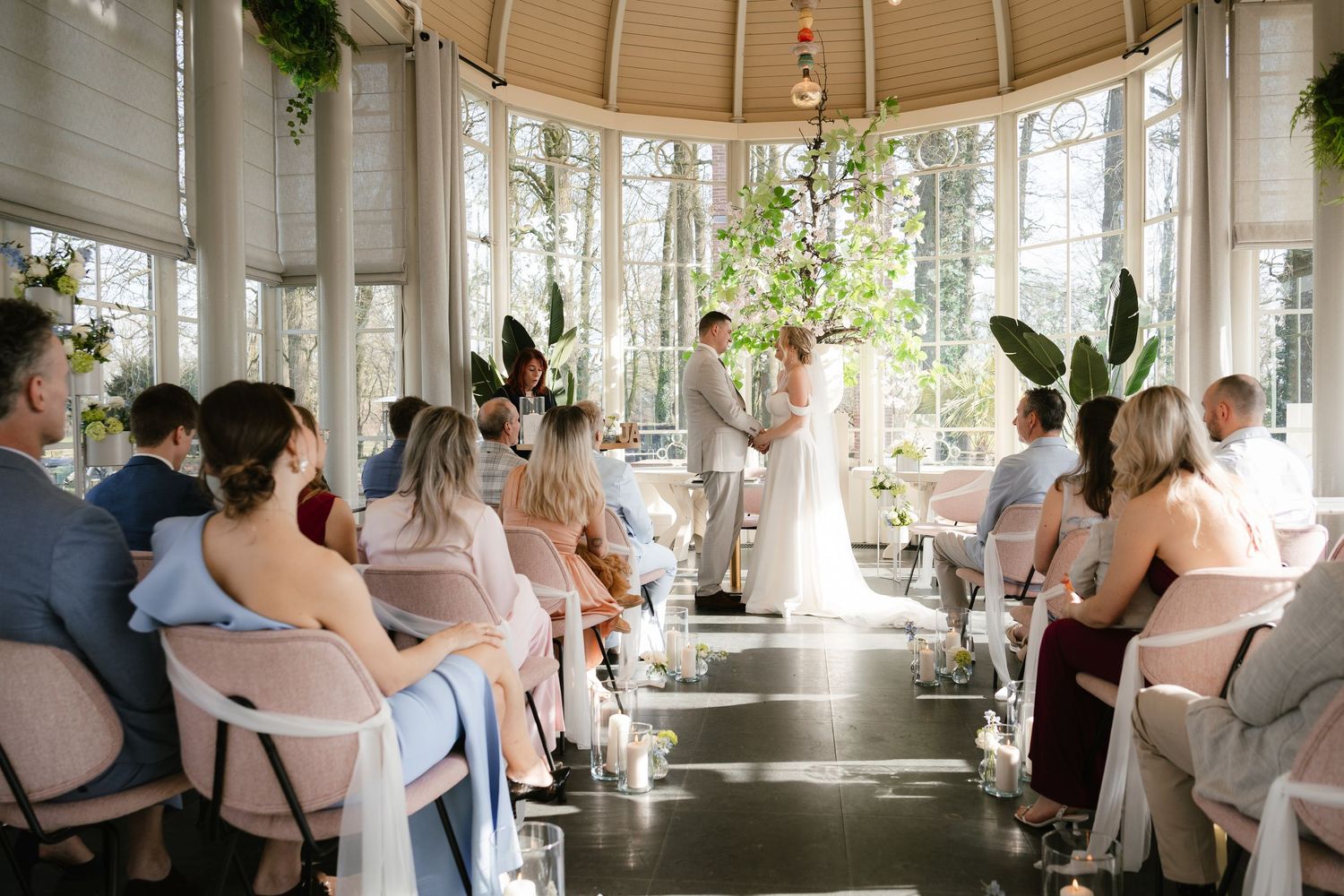 Elegant indoor wedding ceremony in a sunlit glass pavilion with guests seated as couple exchanges vows at the altar.