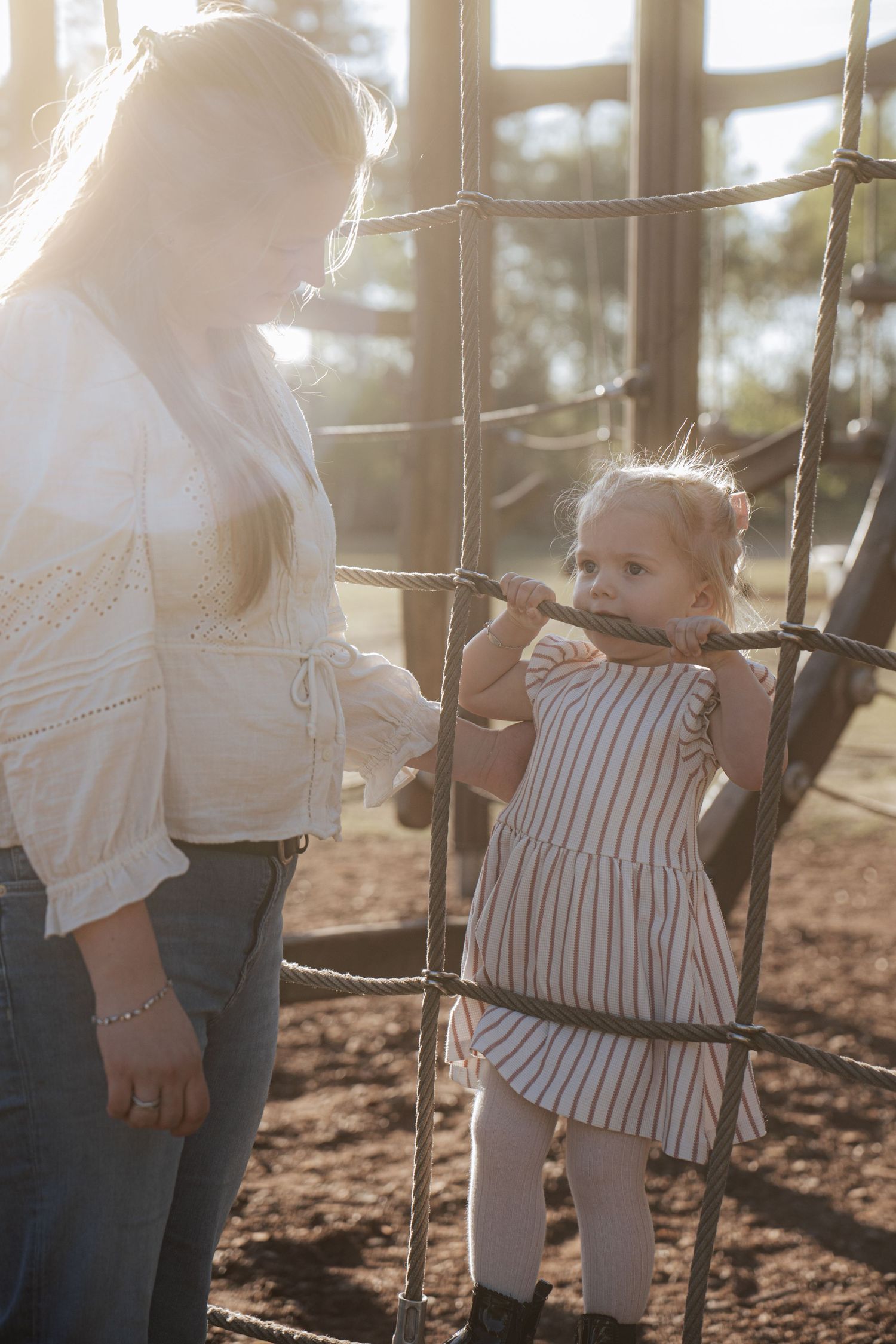A mother watches her toddler daughter play on a rope climbing frame at a sunny playground.