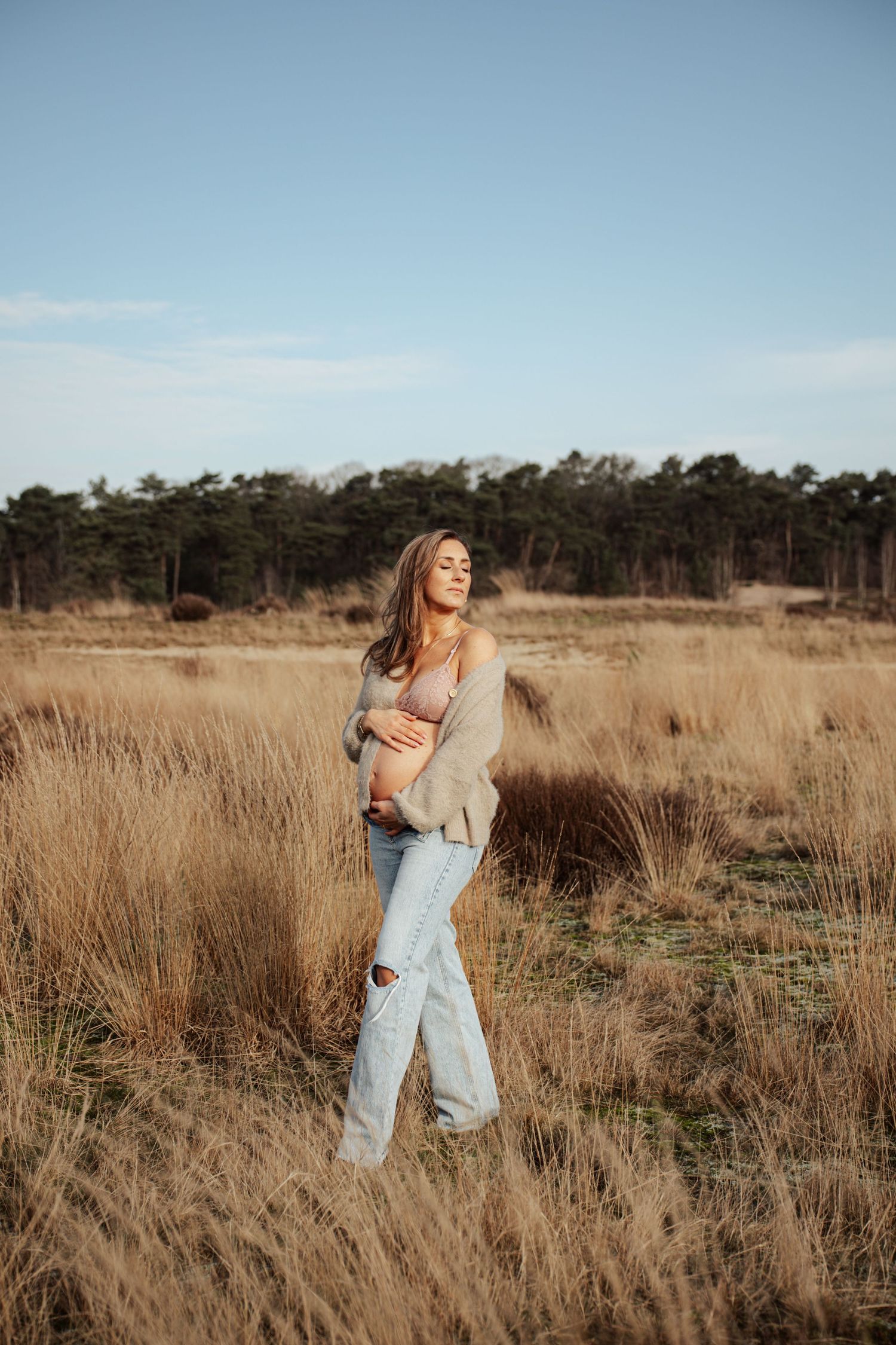 Woman in casual attire stands joyfully in tall golden grass during sunset.