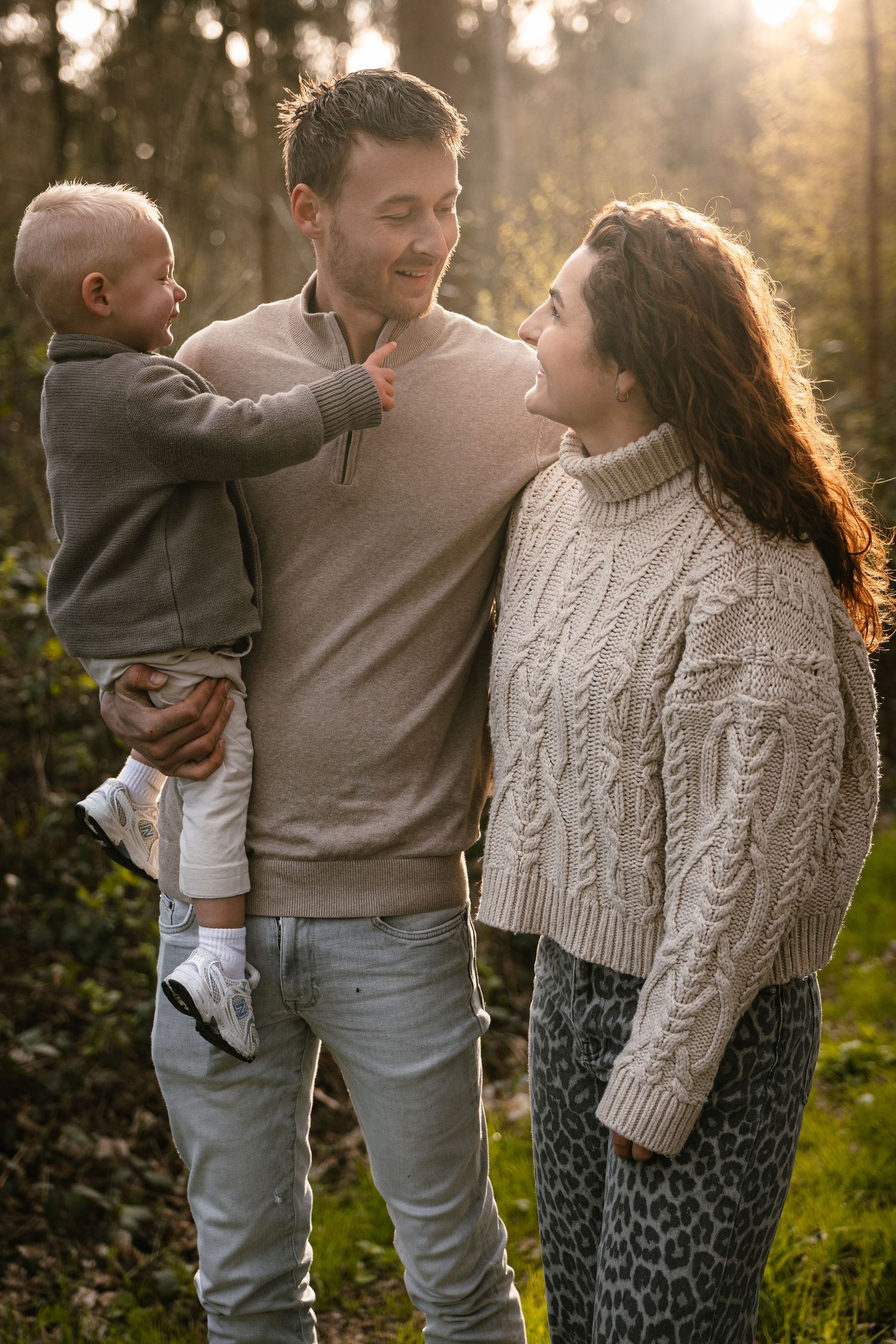 A happy young family with a baby enjoys a sunny outdoor moment in a forest, wearing cozy neutral sweaters.