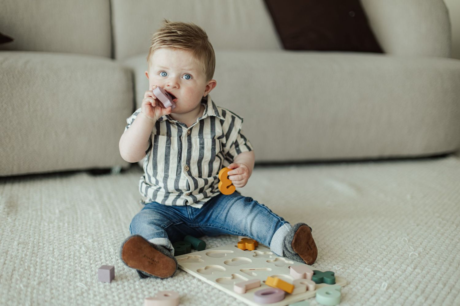 Cute baby boy in striped shirt and jeans sitting on carpet chewing toy near colorful wooden puzzle board.