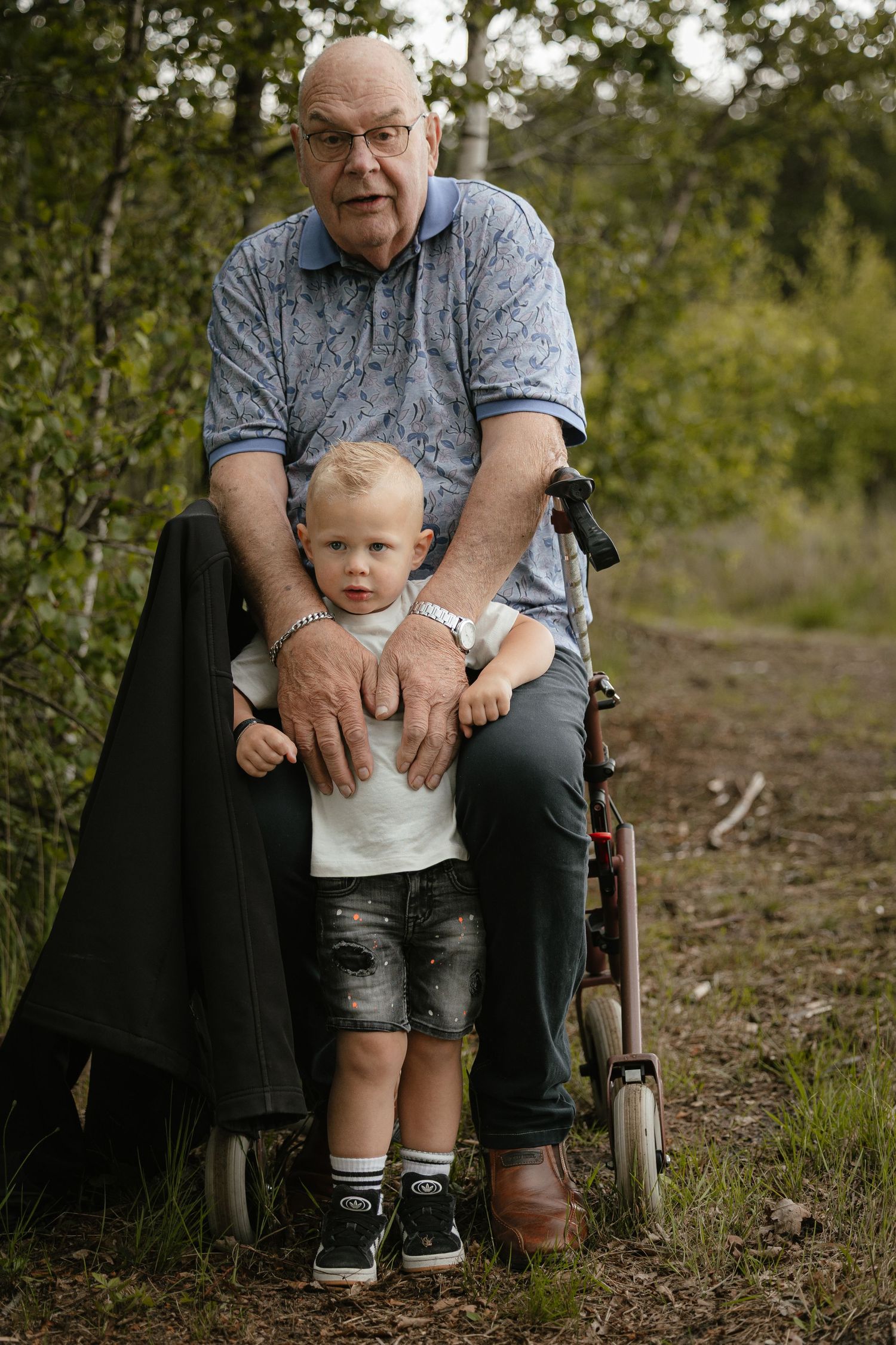 Elderly man in wheelchair with young toddler boy standing between his knees outdoors in a green forest setting.