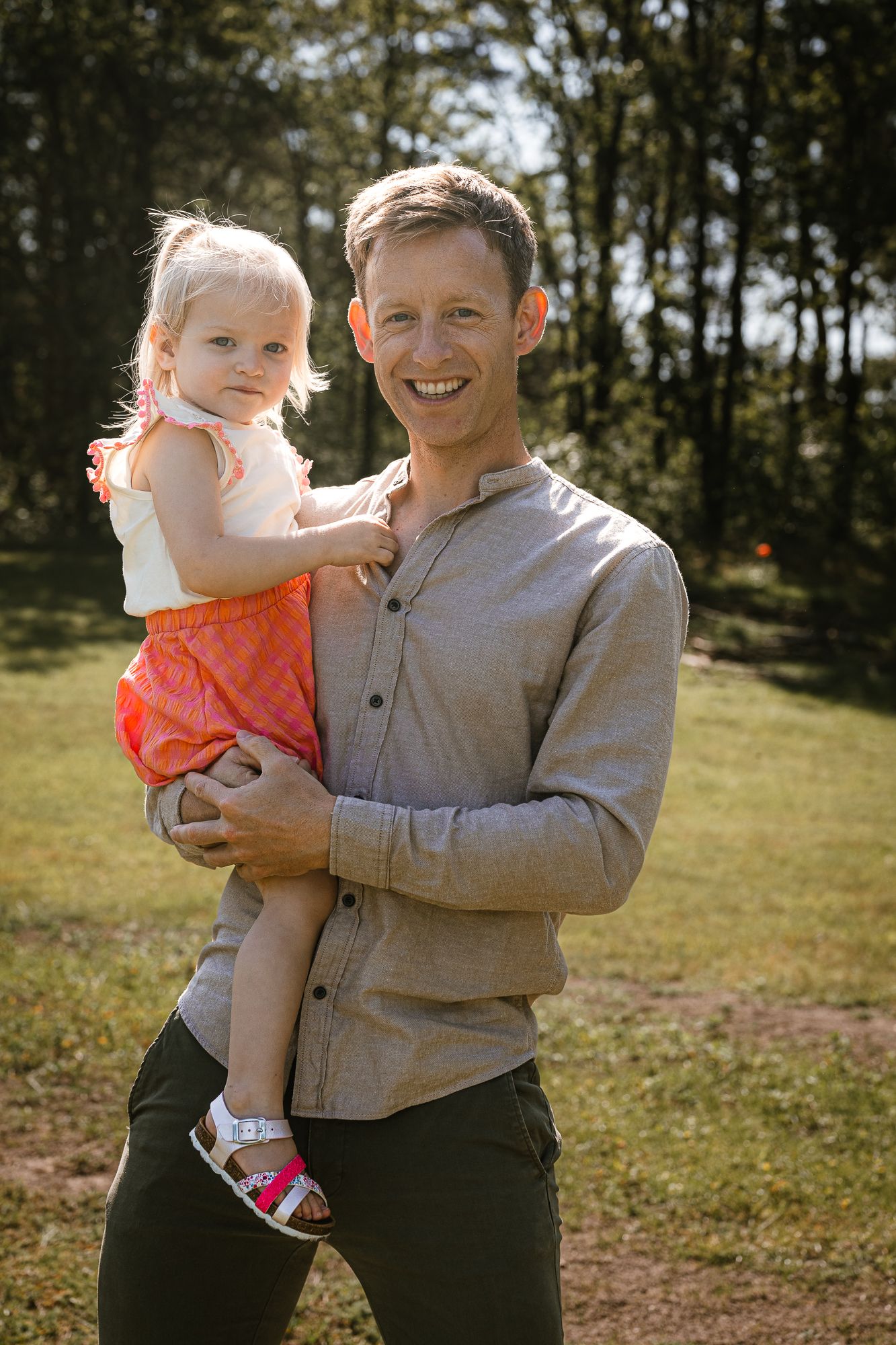 Smiling man holding a toddler girl in a pink dress outdoors in a sunny green park setting.