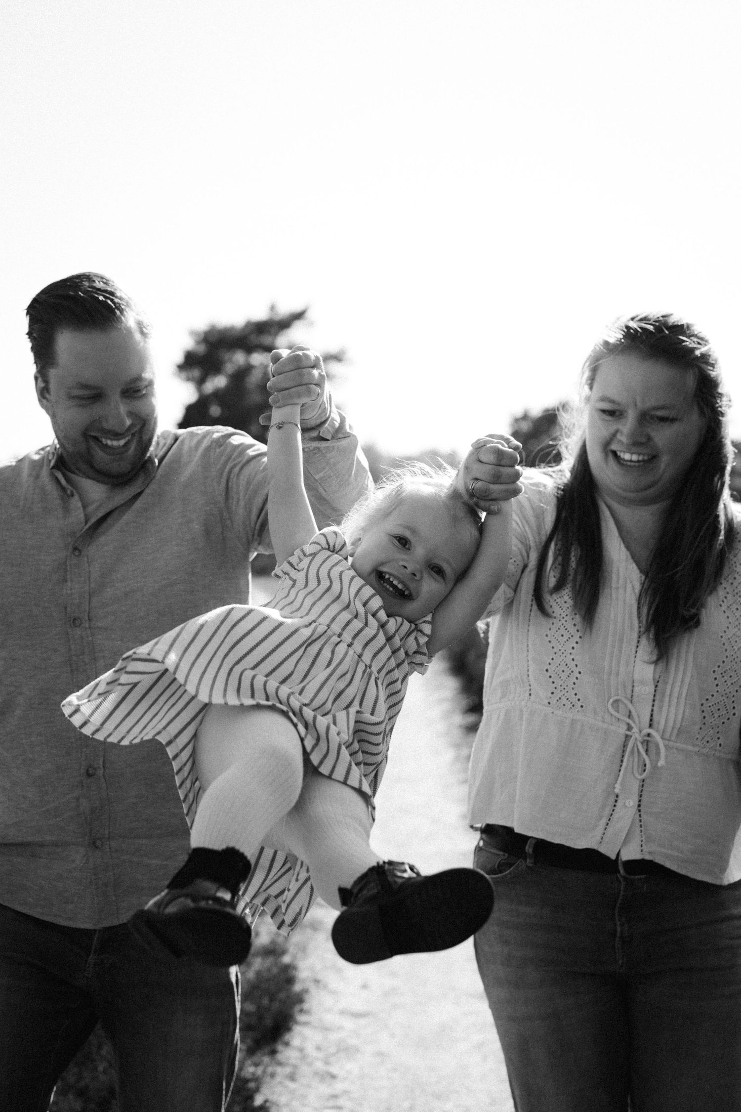 Black and white photo of happy parents swinging their laughing toddler by the hands outdoors.