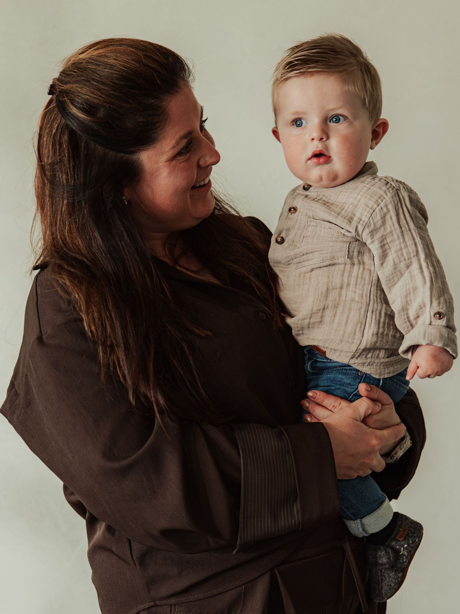 A woman with long brown hair holds a smiling baby boy in a beige shirt and jeans against a light background.