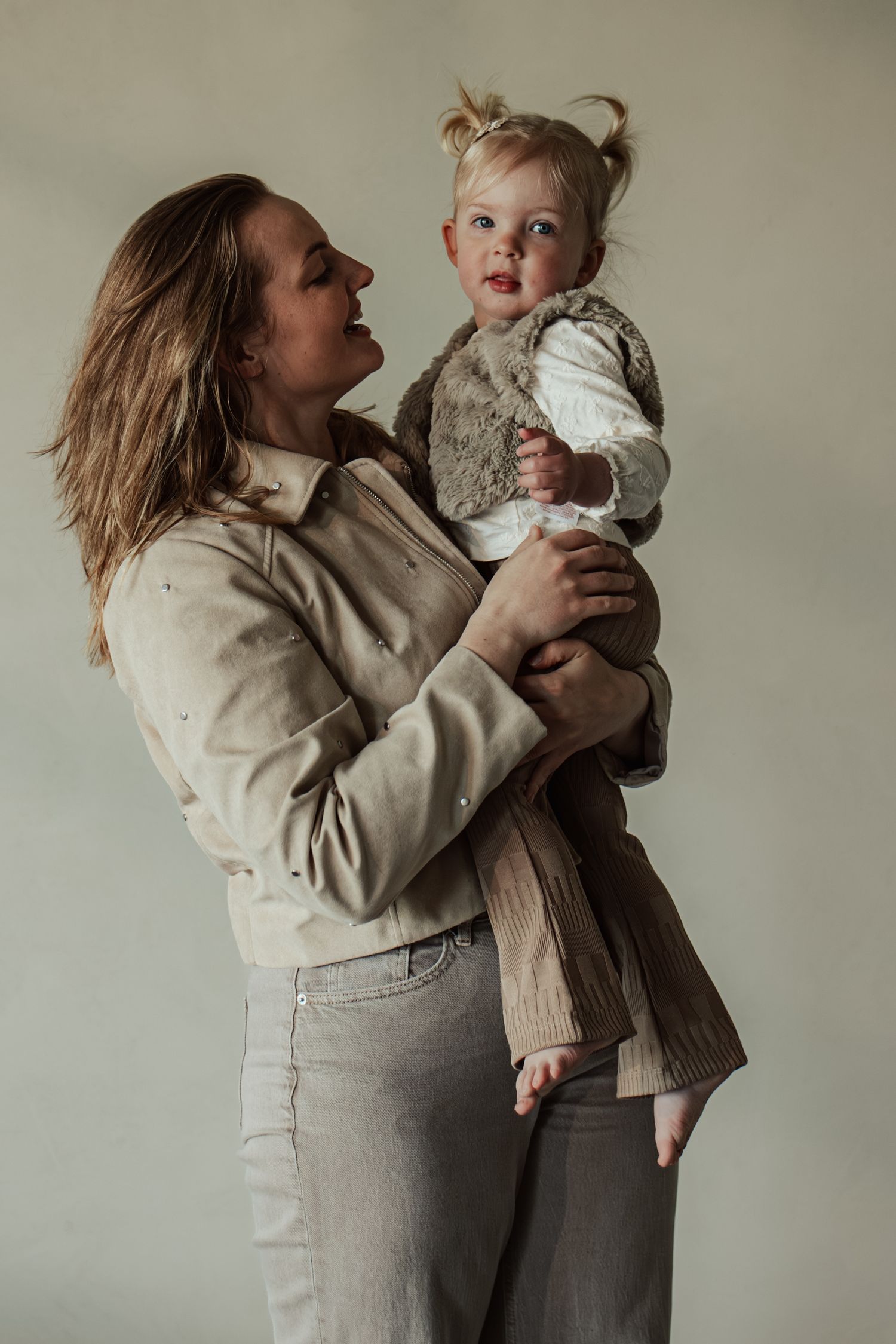 A woman in a beige jacket holds a smiling toddler with pigtails against a neutral background.