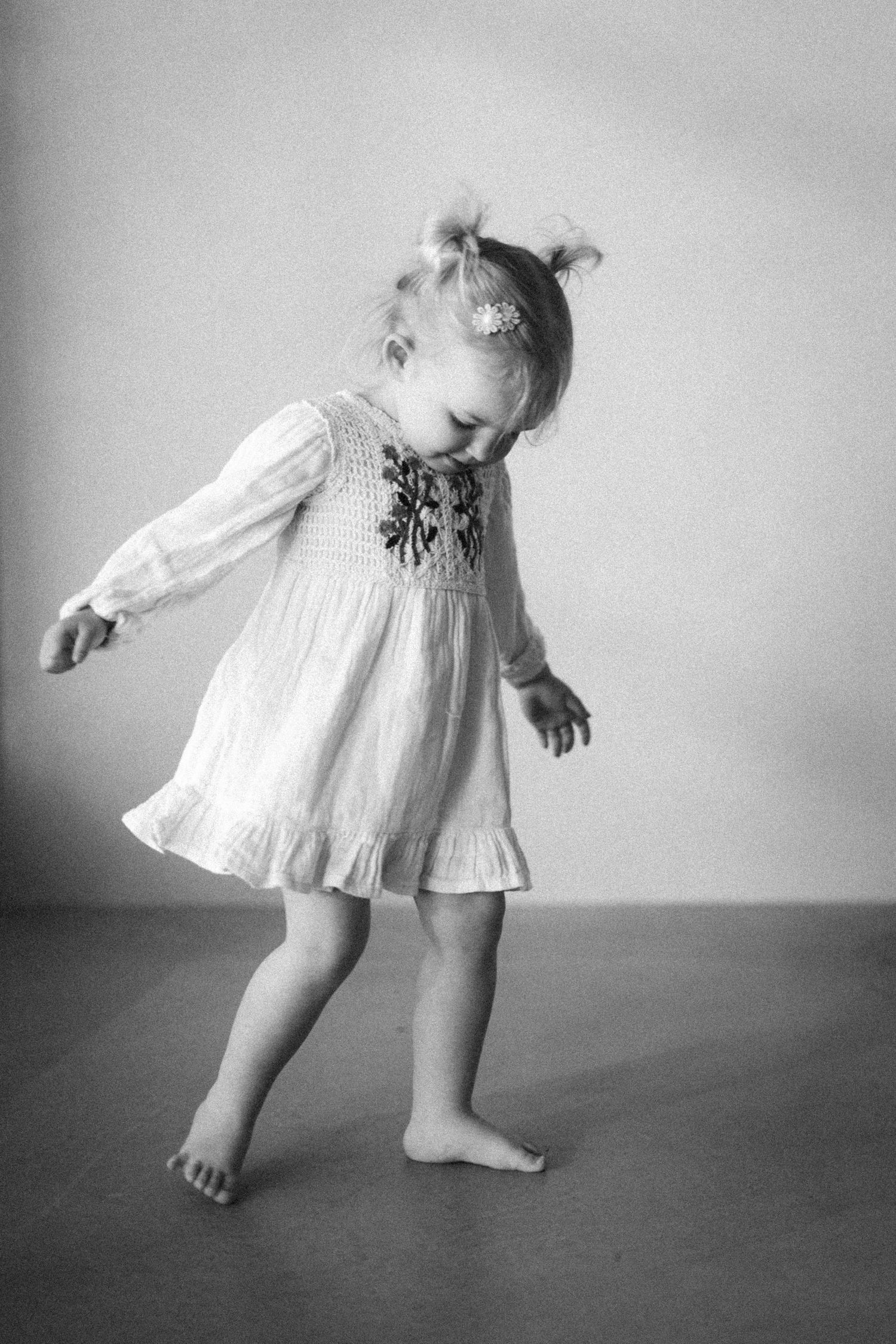 Black and white photo of a toddler girl in a floral dress dancing barefoot, with pigtails and a flower hair clip.