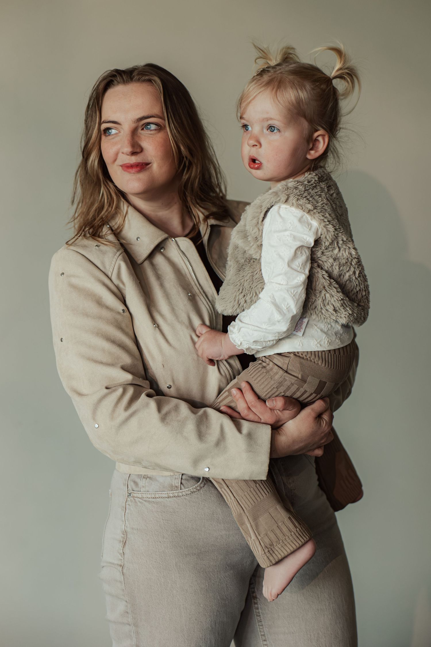 A young woman in beige jacket and jeans holds a toddler girl in a fur vest against a neutral background.