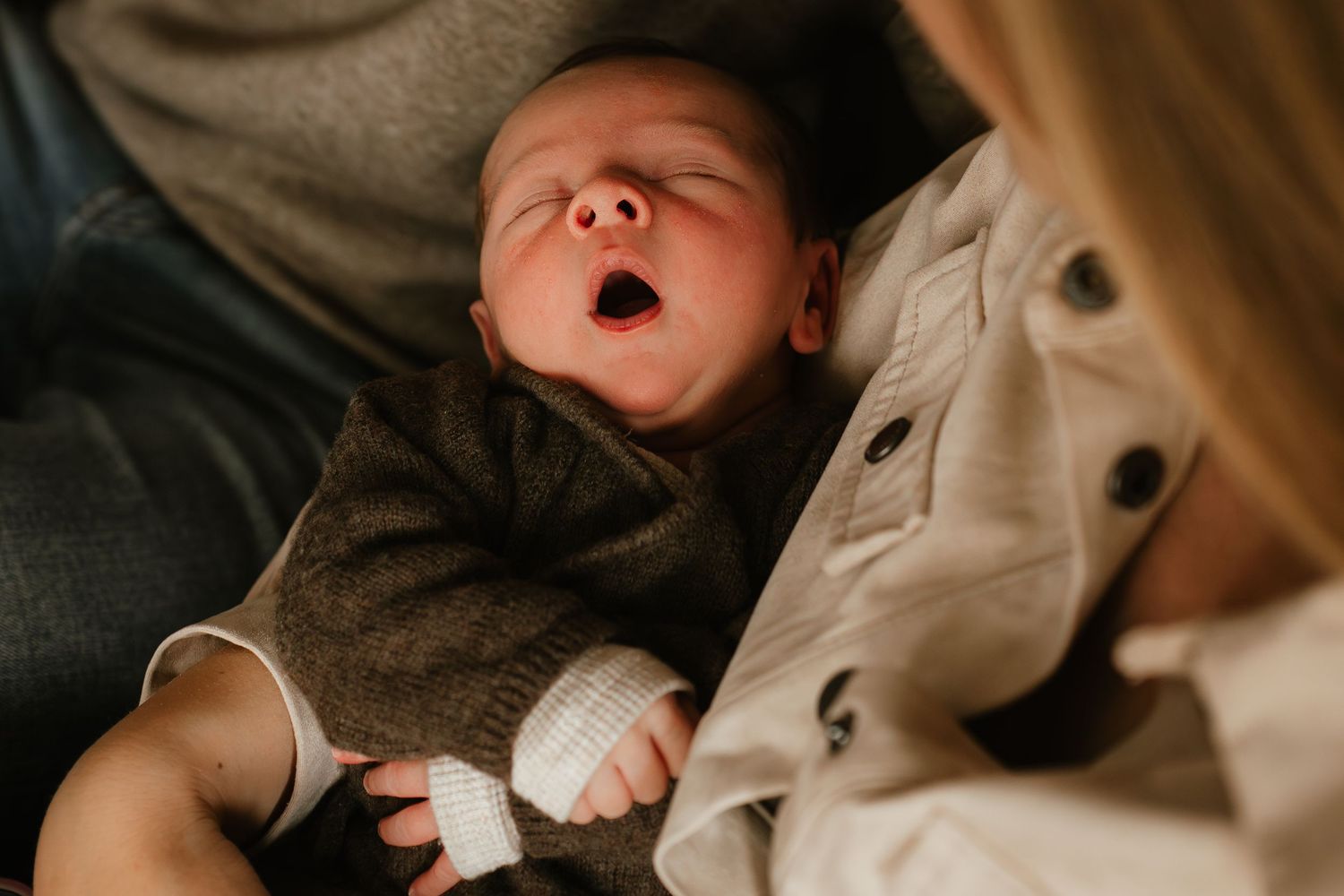 Newborn baby yawning while being held by mother, wearing a brown knit outfit during a lifestyle newborn photo session.