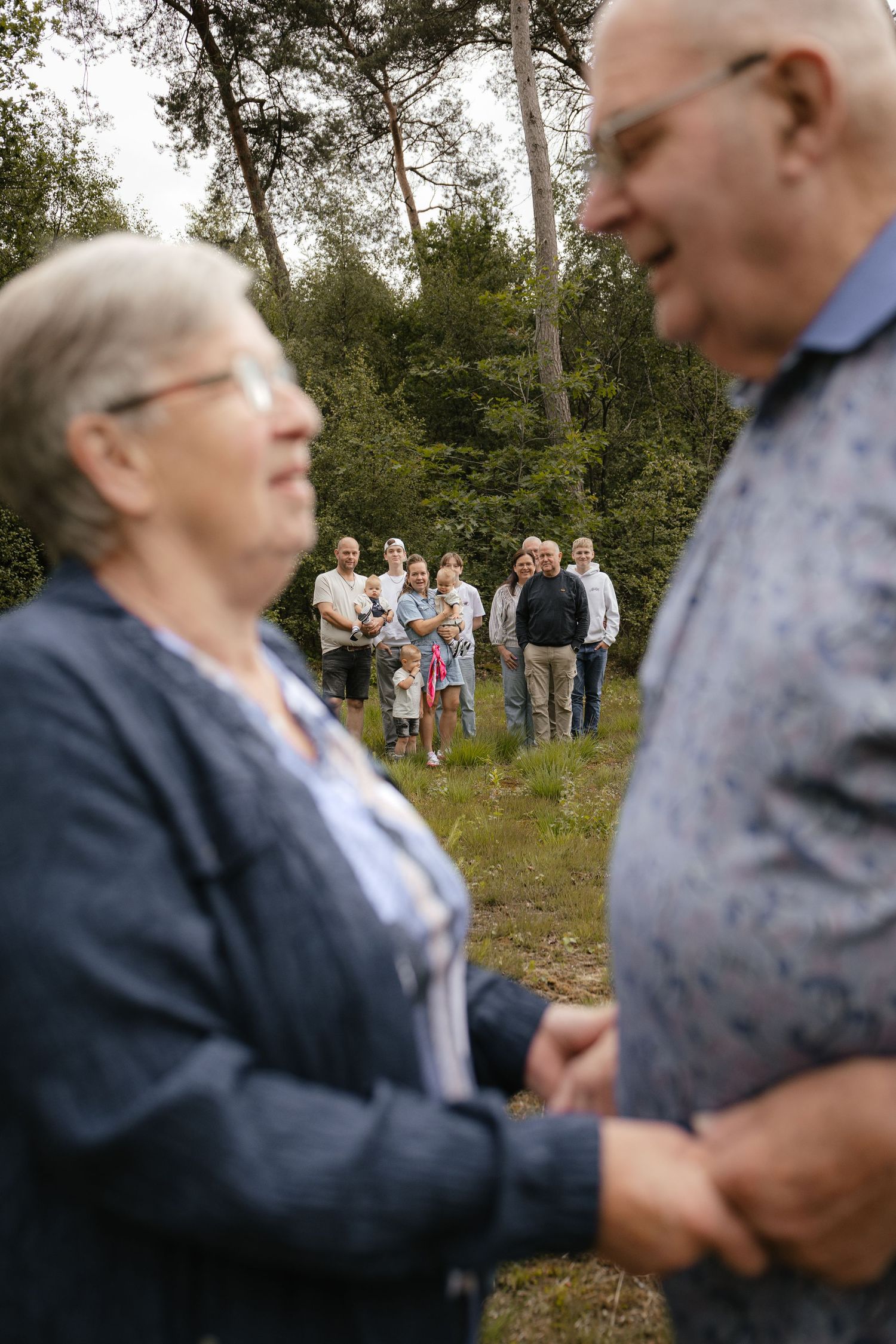 Elderly couple holding hands outdoors while a group of family members stands in the background near trees.
