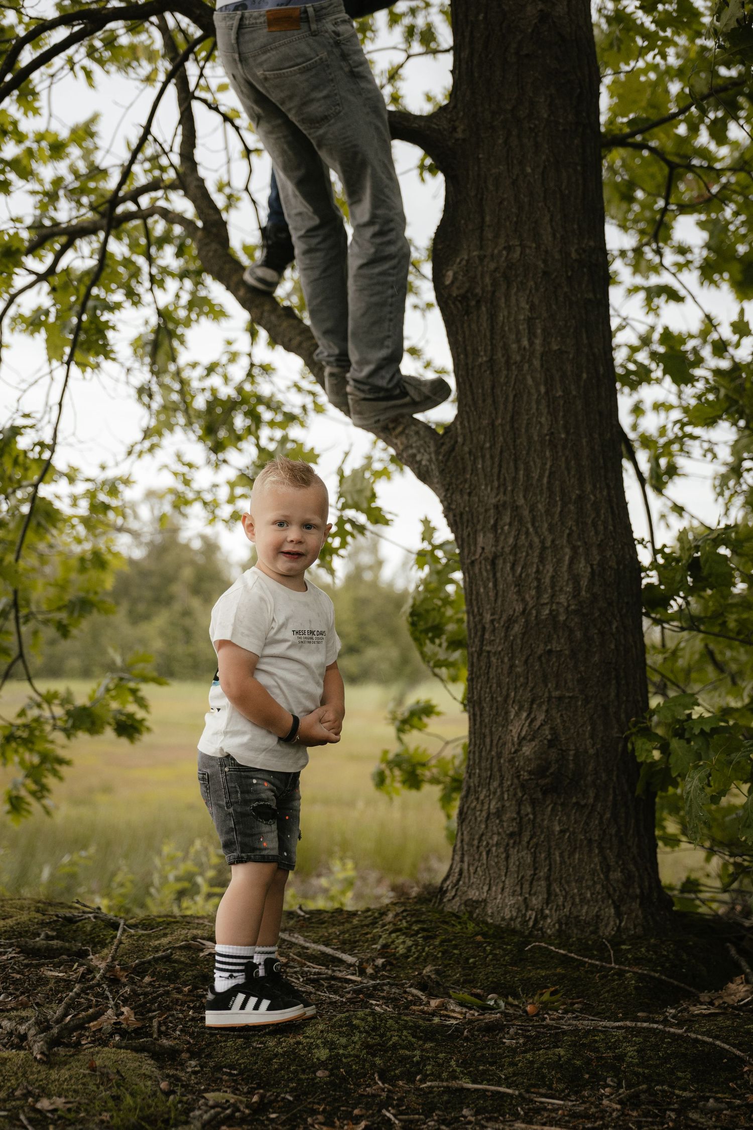 Young boy in white t-shirt and shorts stands smiling by a tree while another person climbs branches above him.