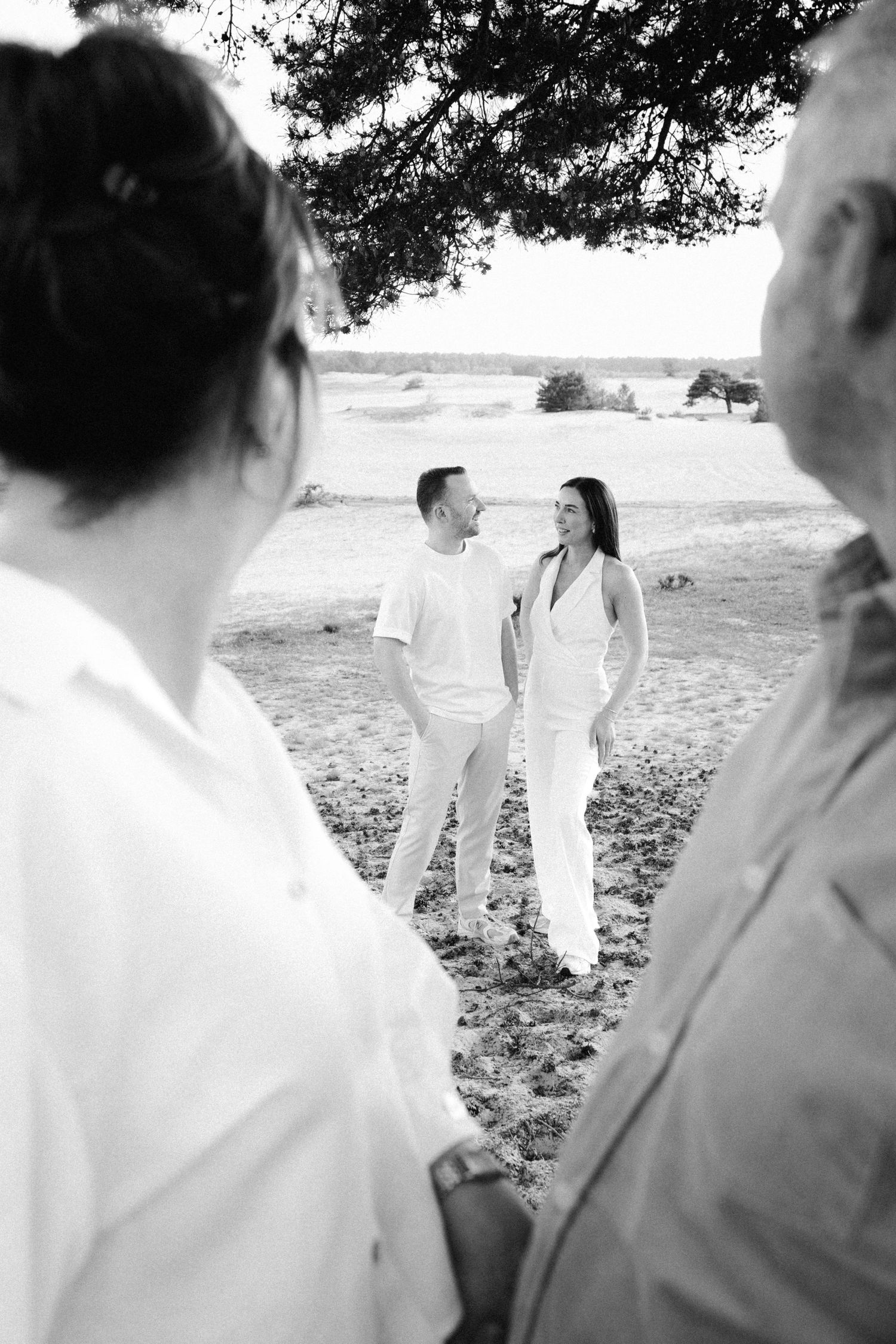 Black and white photo of a couple in white attire standing in a field, smiling at each other, observed by guests.