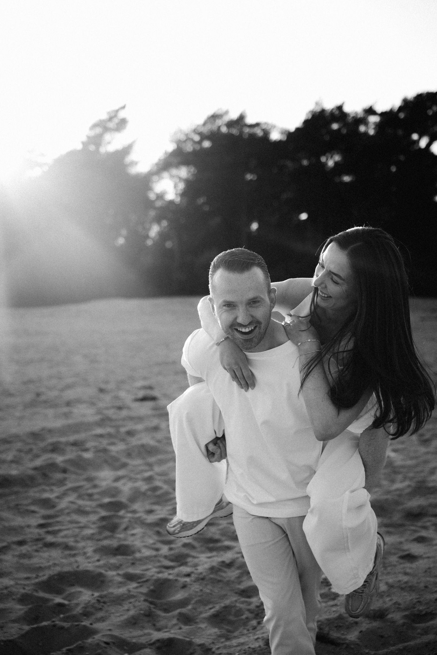 Black and white photo of man giving woman a piggyback ride on sandy beach.