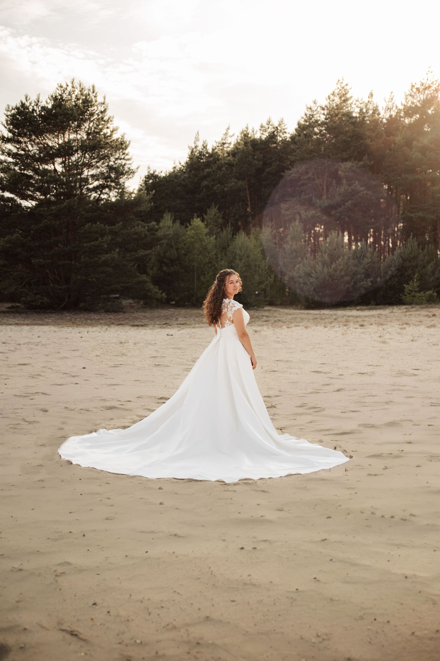 Bride in flowing white wedding dress walking across sandy dunes at sunset.