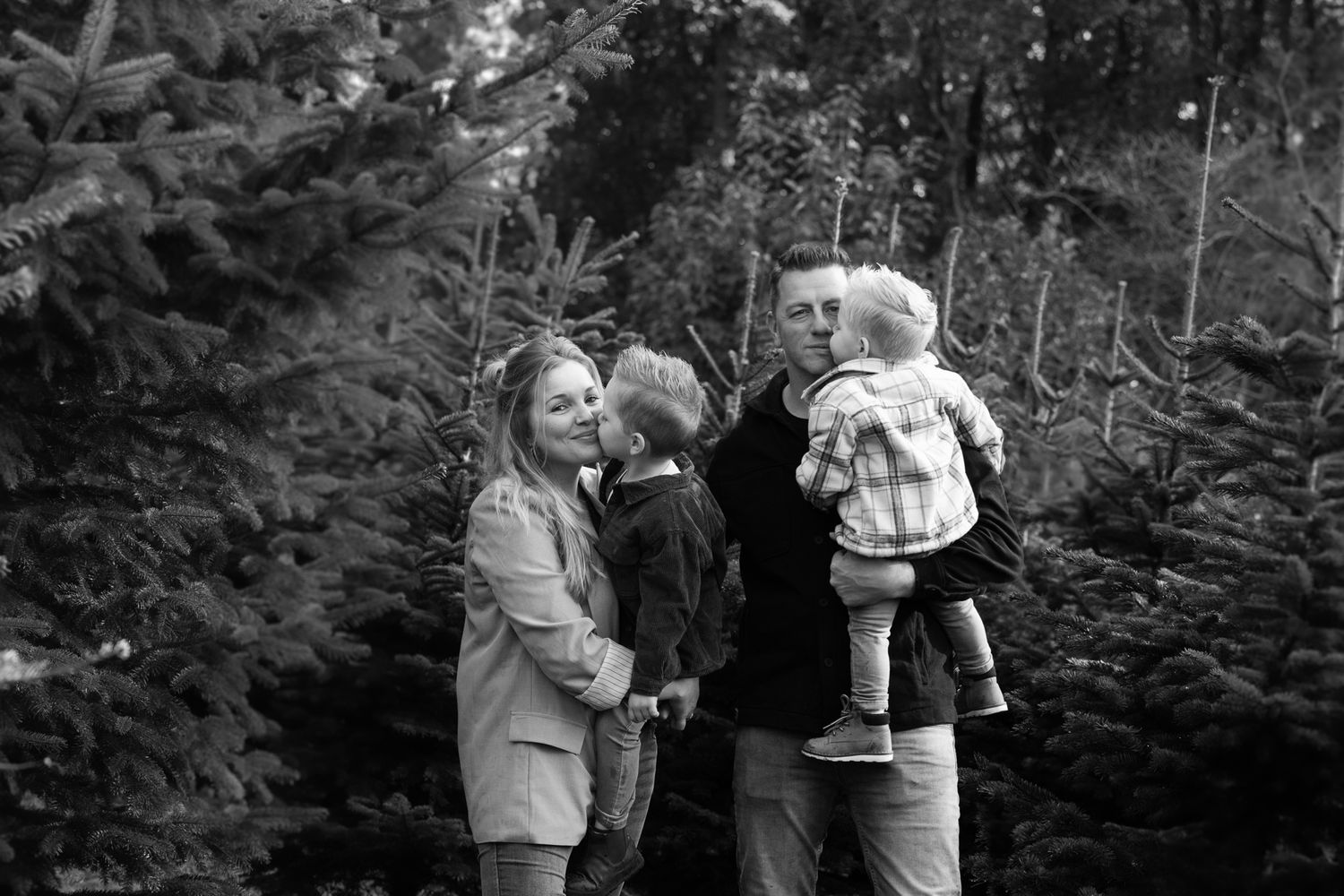Family enjoying a playful moment among evergreen trees in black and white.